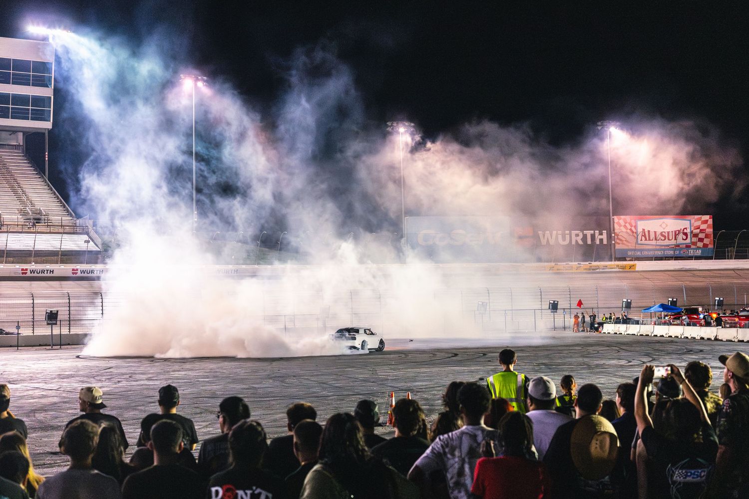 White car engulfed in tire smoke during a nighttime burnout show at a racing venue.