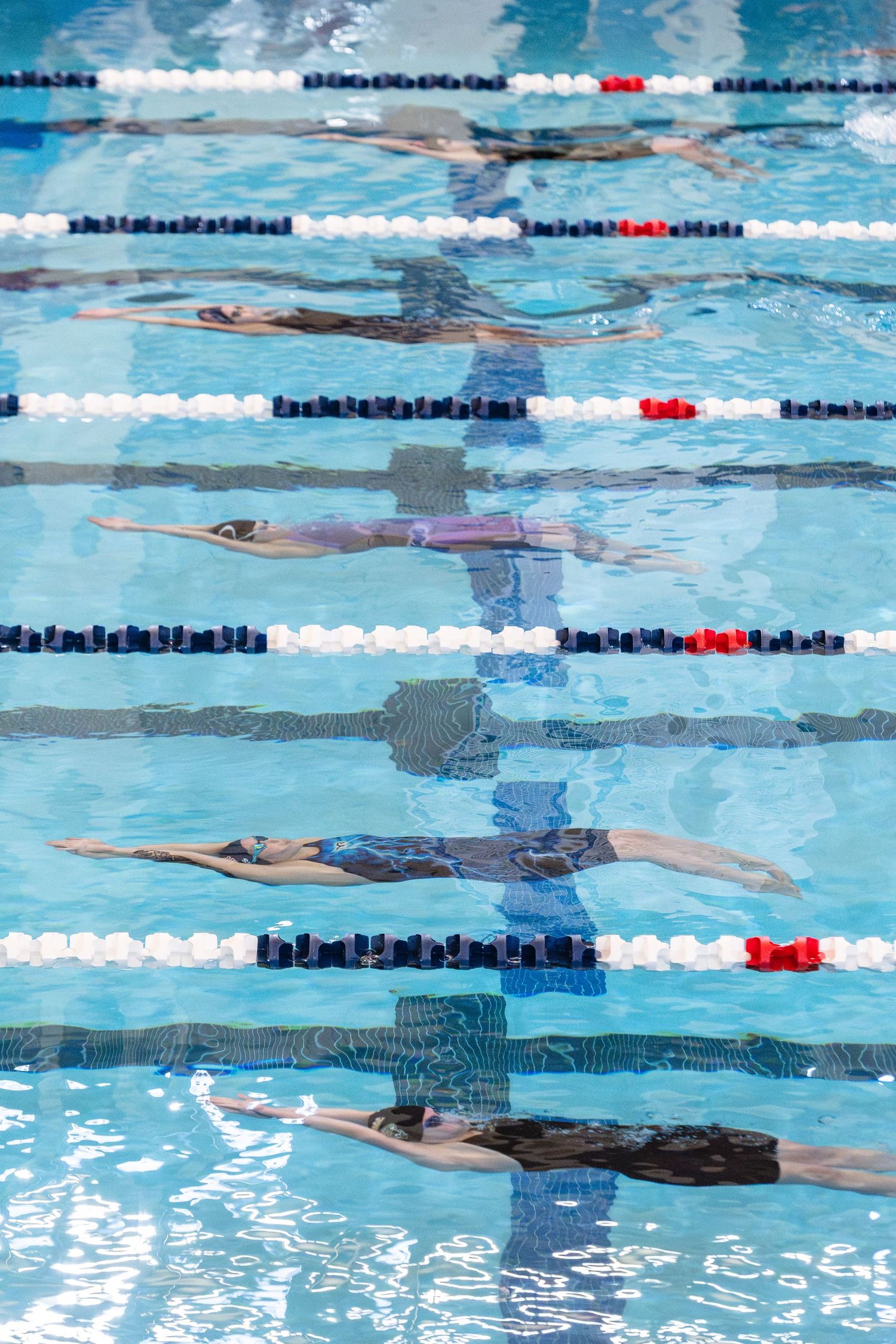 Competitive swimmers race in multiple lanes of a bright blue indoor swimming pool during a swim meet.
