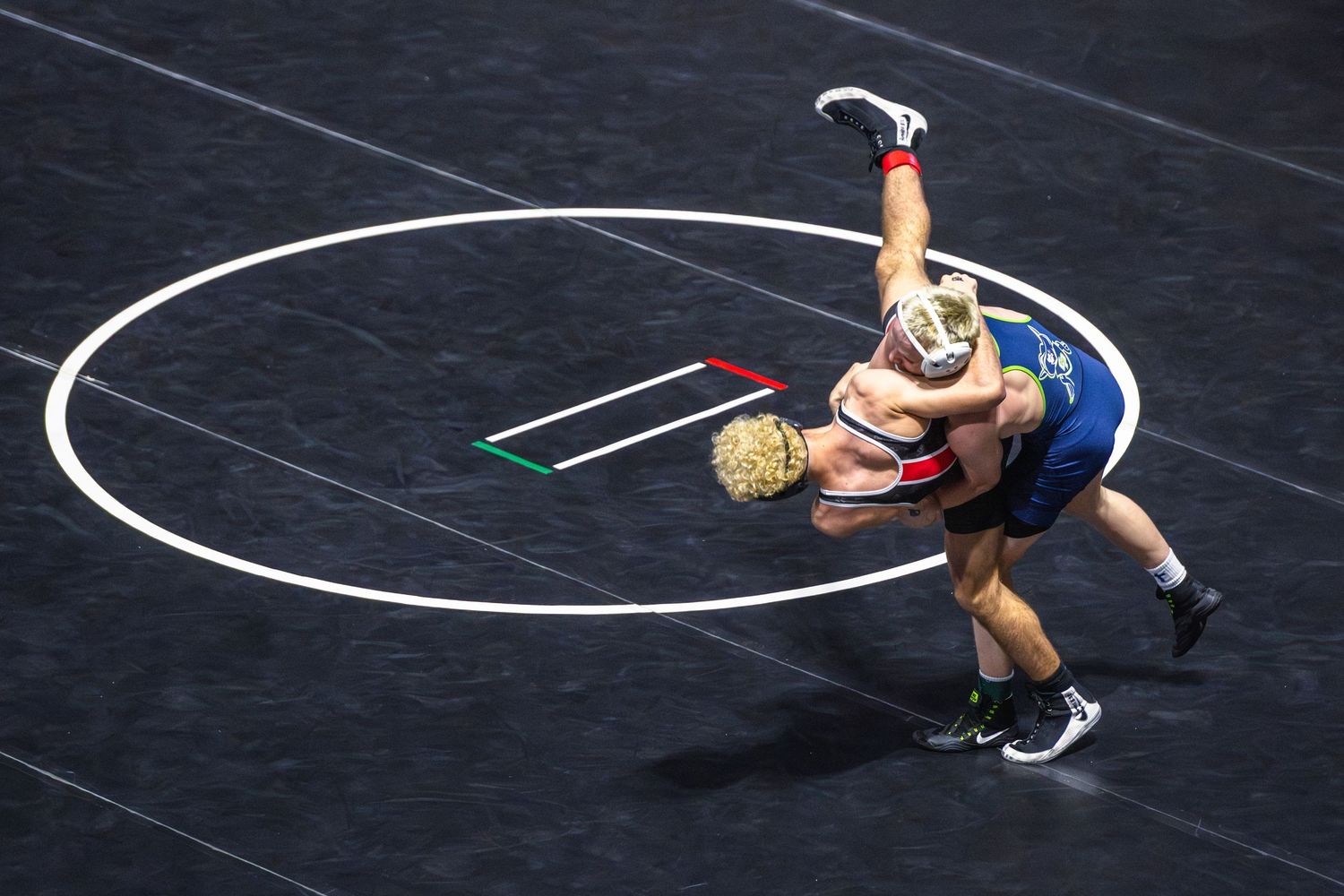 Two wrestlers compete on a mat, one lifting the other overhead during an intense wrestling match.