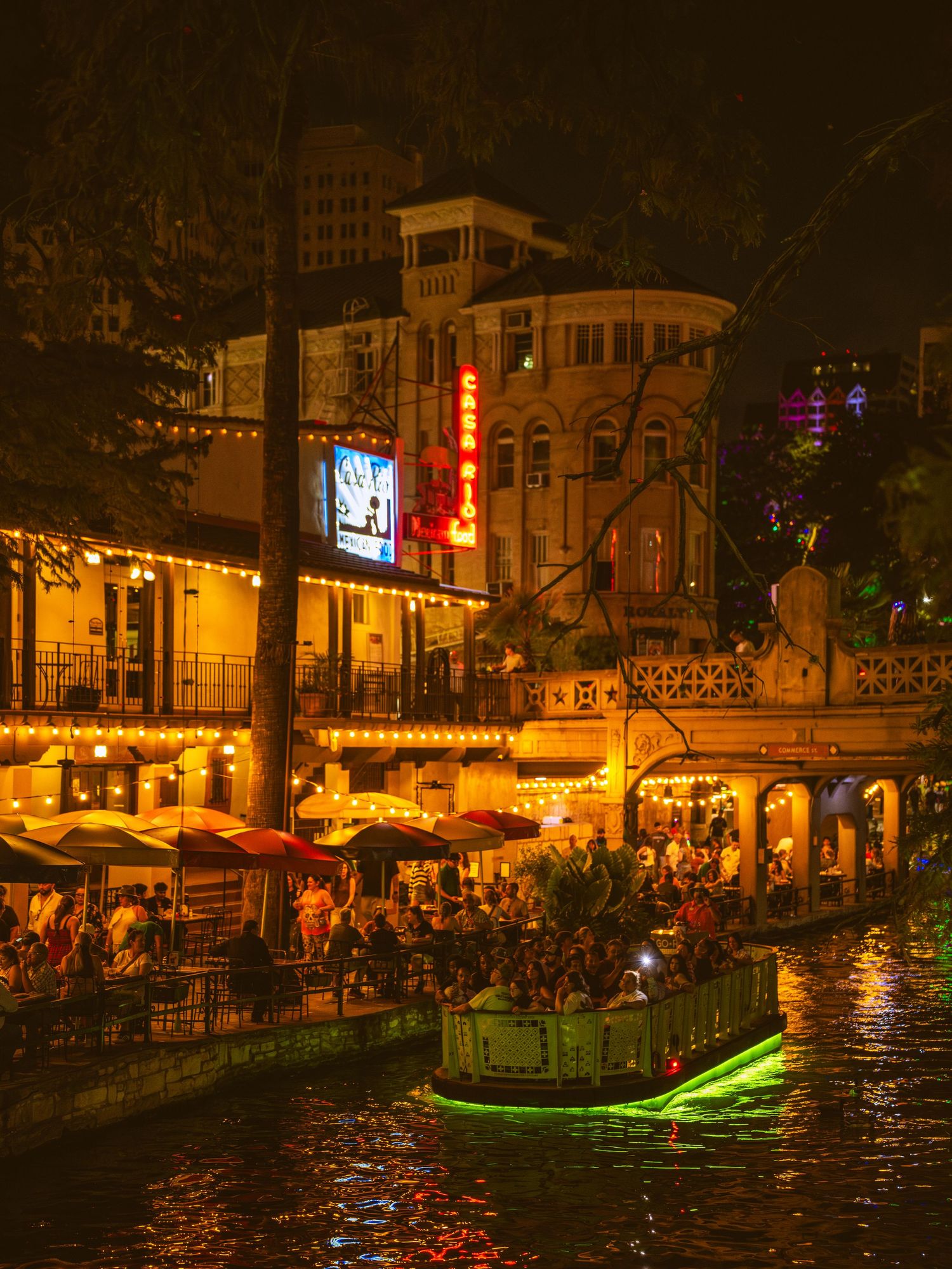 Illuminated San Antonio River Walk at night with glowing boat and bustling waterfront restaurants.