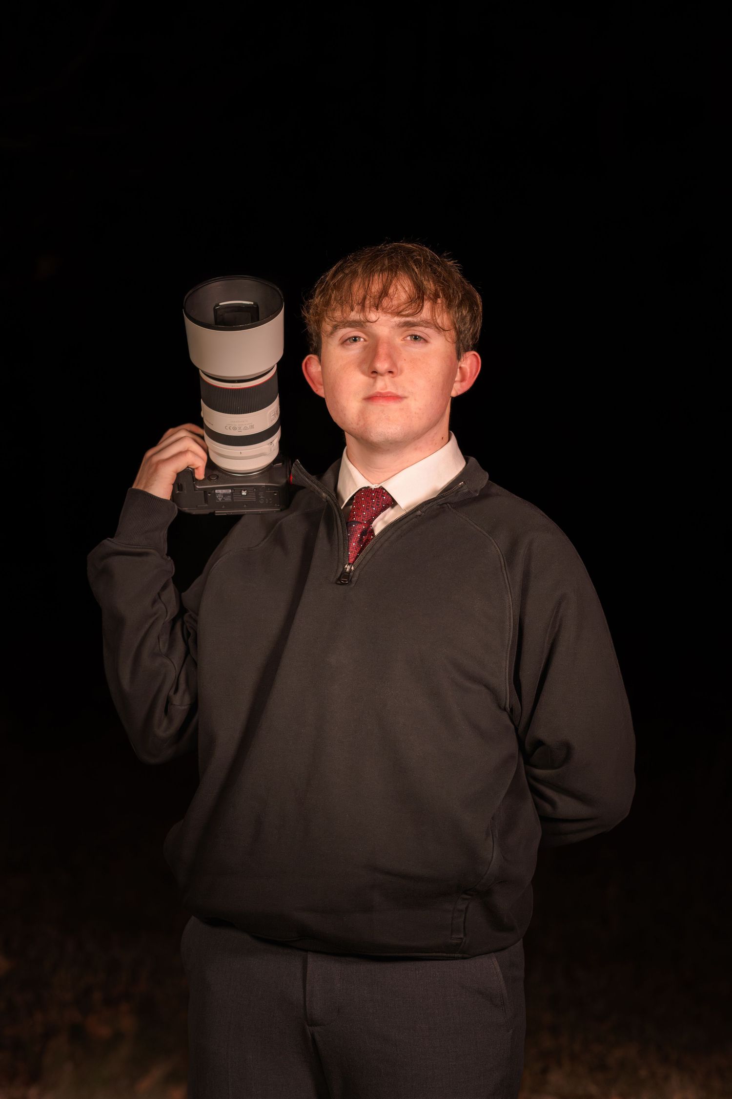 Young man holding a large Canon telephoto lens camera on his shoulder against a dark background.