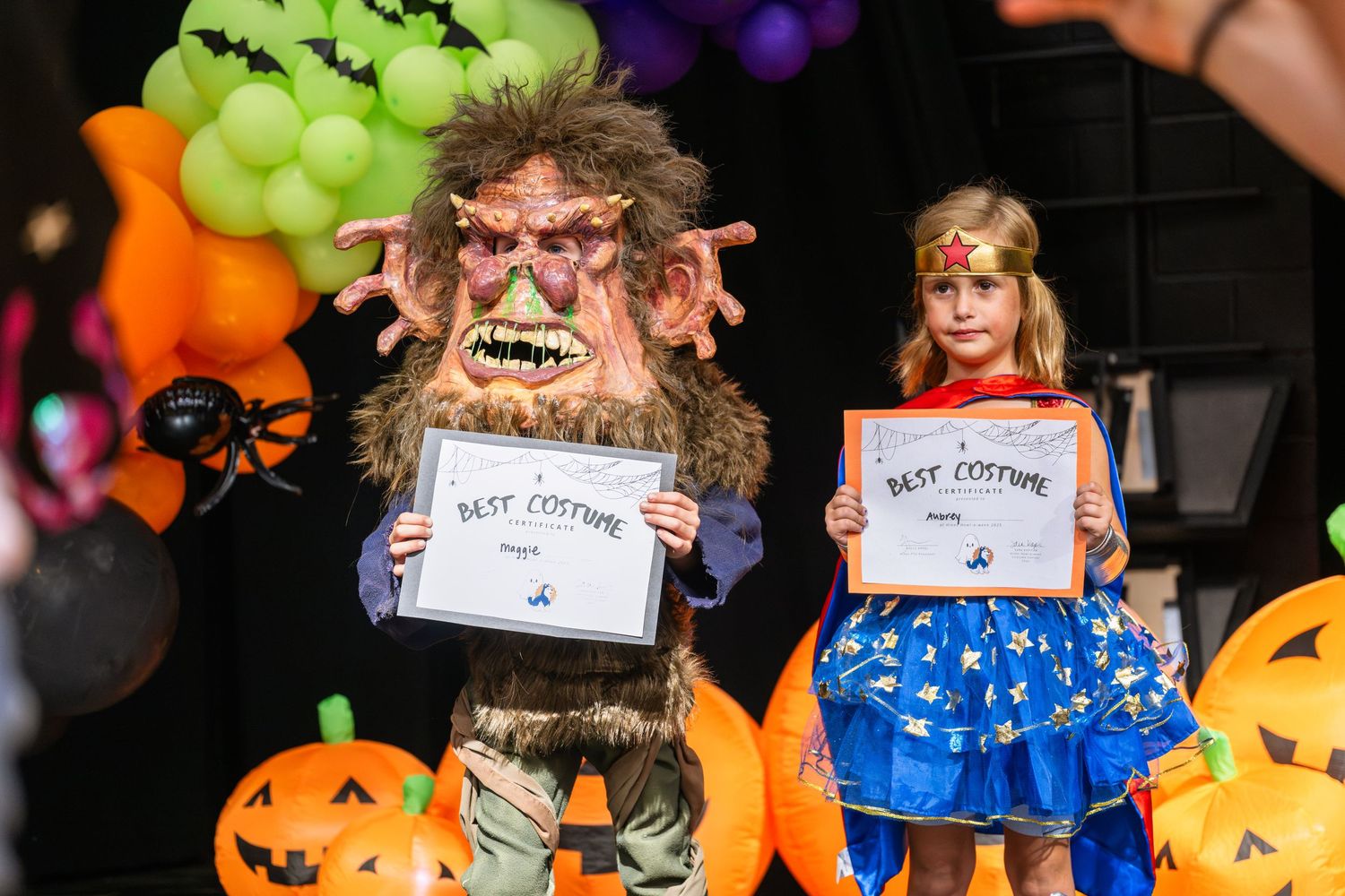 Two Halloween costume contest winners holding Best Costume certificates, a troll and a fairy princess, on a decorated stage.
