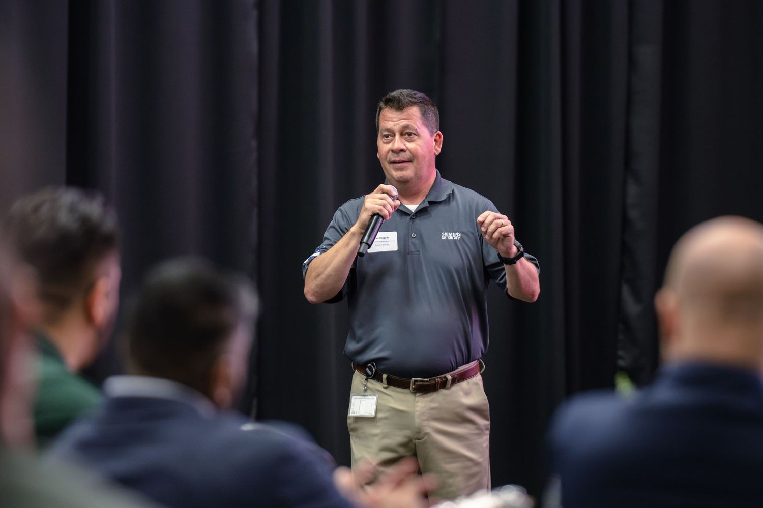 Man in gray polo shirt speaks to audience at corporate event, holding microphone on stage with black curtain backdrop.