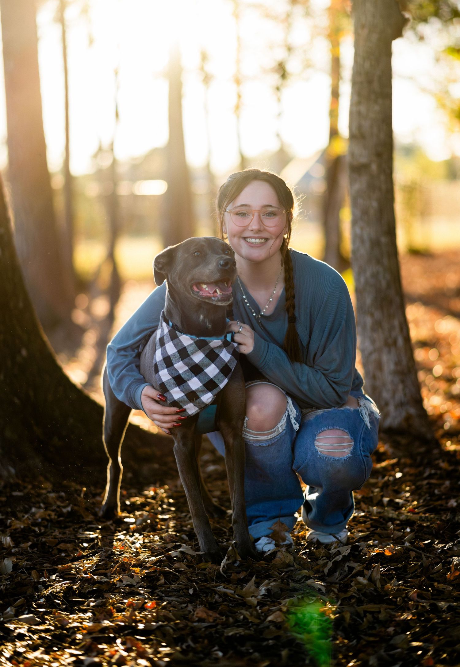 Young woman crouching with a dark brown dog wearing a plaid bandana in a sunlit autumn forest.