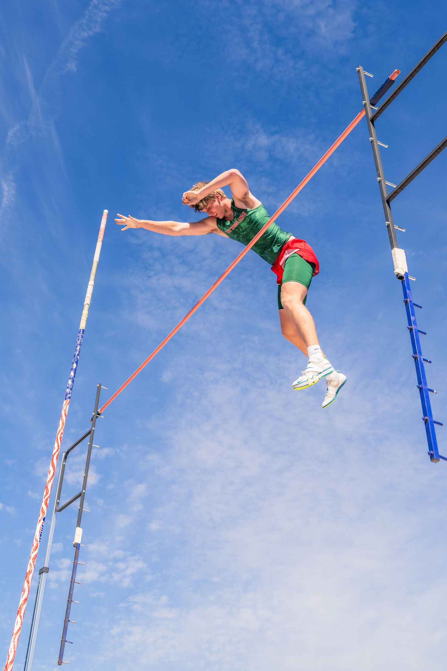 Male athlete performs pole vault against bright blue sky, body arched over the red bar during track competition.