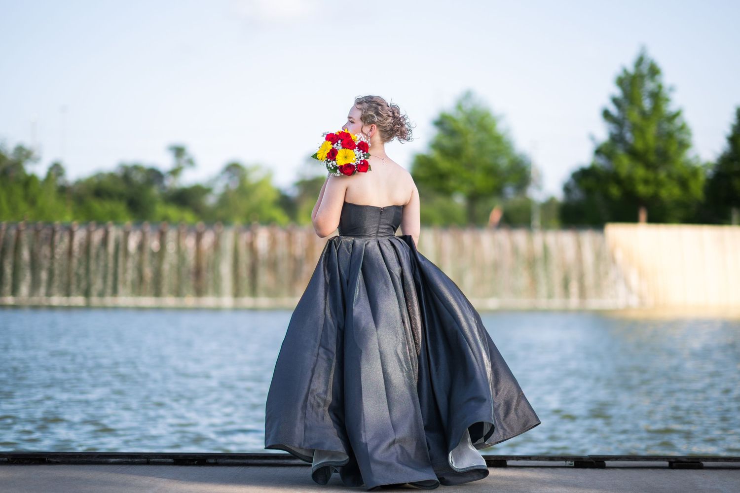 Young lady in black strapless ball gown holding colorful bouquet by waterfront outdoor venue.
