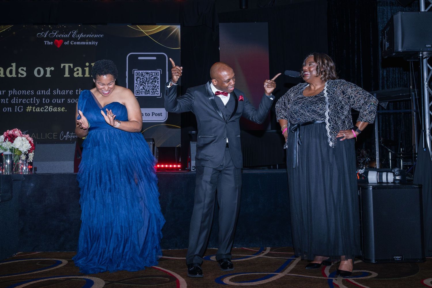 Three people dance and celebrate on stage at a formal gala event with a presentation screen behind them.