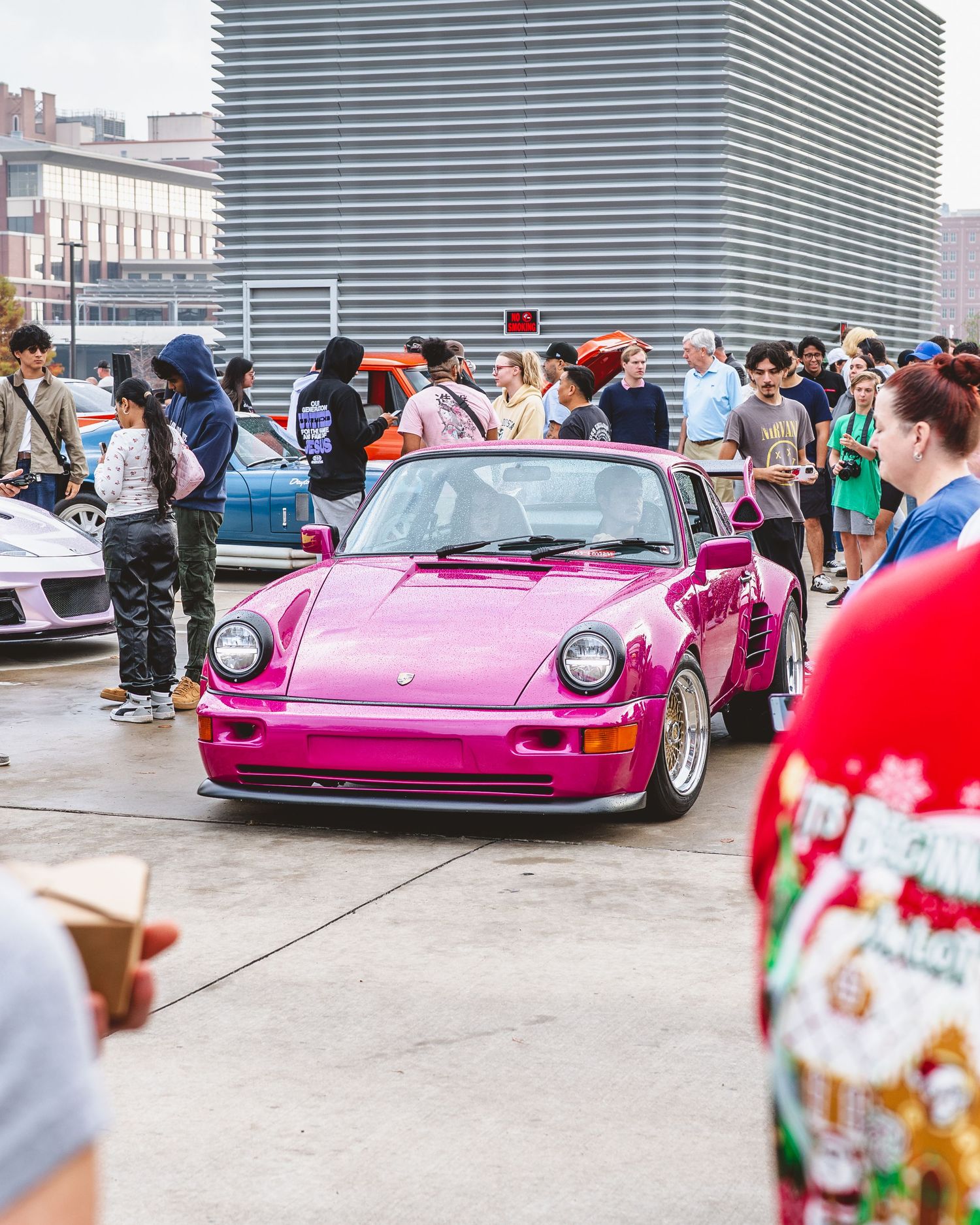 Bright pink Porsche 911 RWB on display at an outdoor car meet with crowds of enthusiasts.