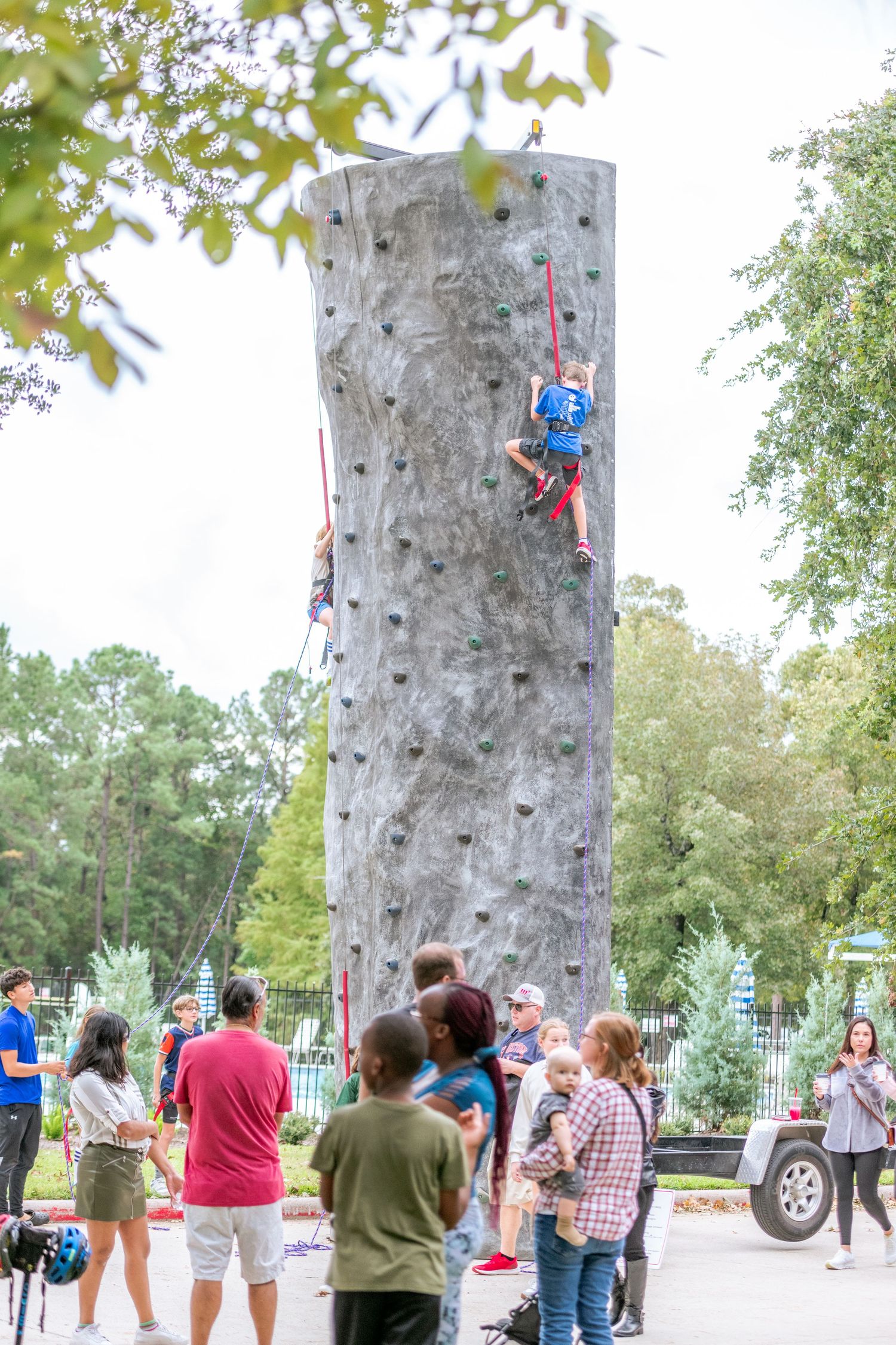 People watching climbers scale a tall outdoor rock climbing wall surrounded by trees at a park event.