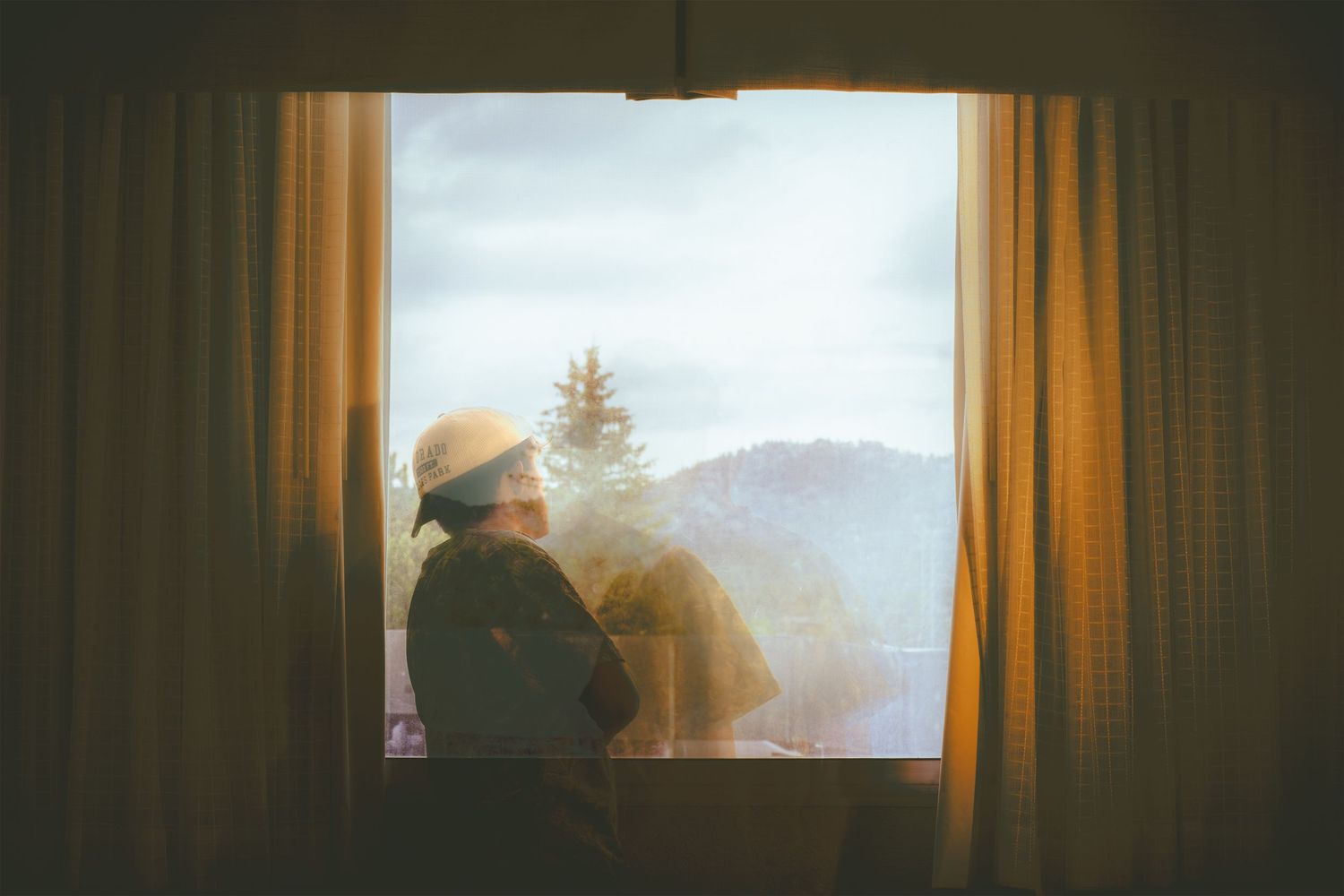A person wearing a hat sits by an open window gazing at a scenic mountain and tree landscape in soft warm light.