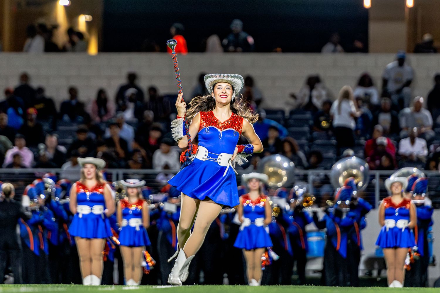Majorette in blue costume twirls baton while leading a marching band performance at a football game.