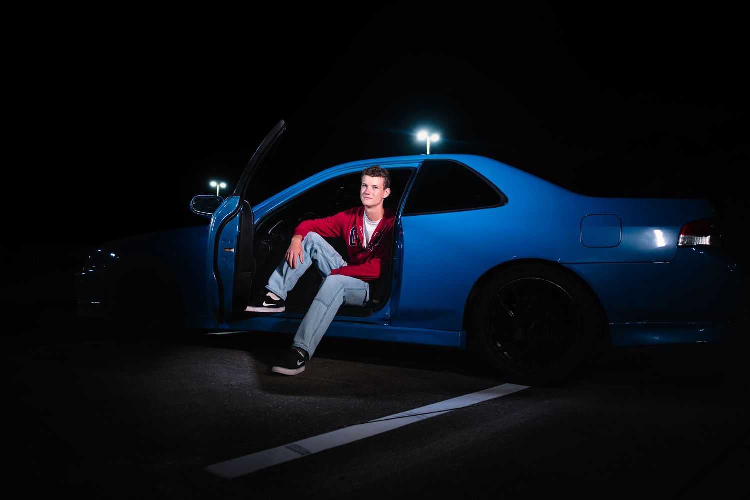 Young man sitting in open car door of blue sports car at night with dramatic lighting.