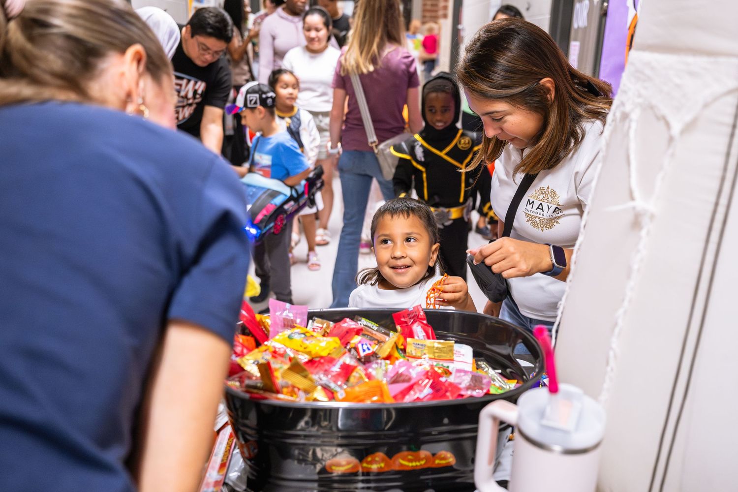 A smiling child reaches for colorful candies at a festive outdoor market stall surrounded by people.