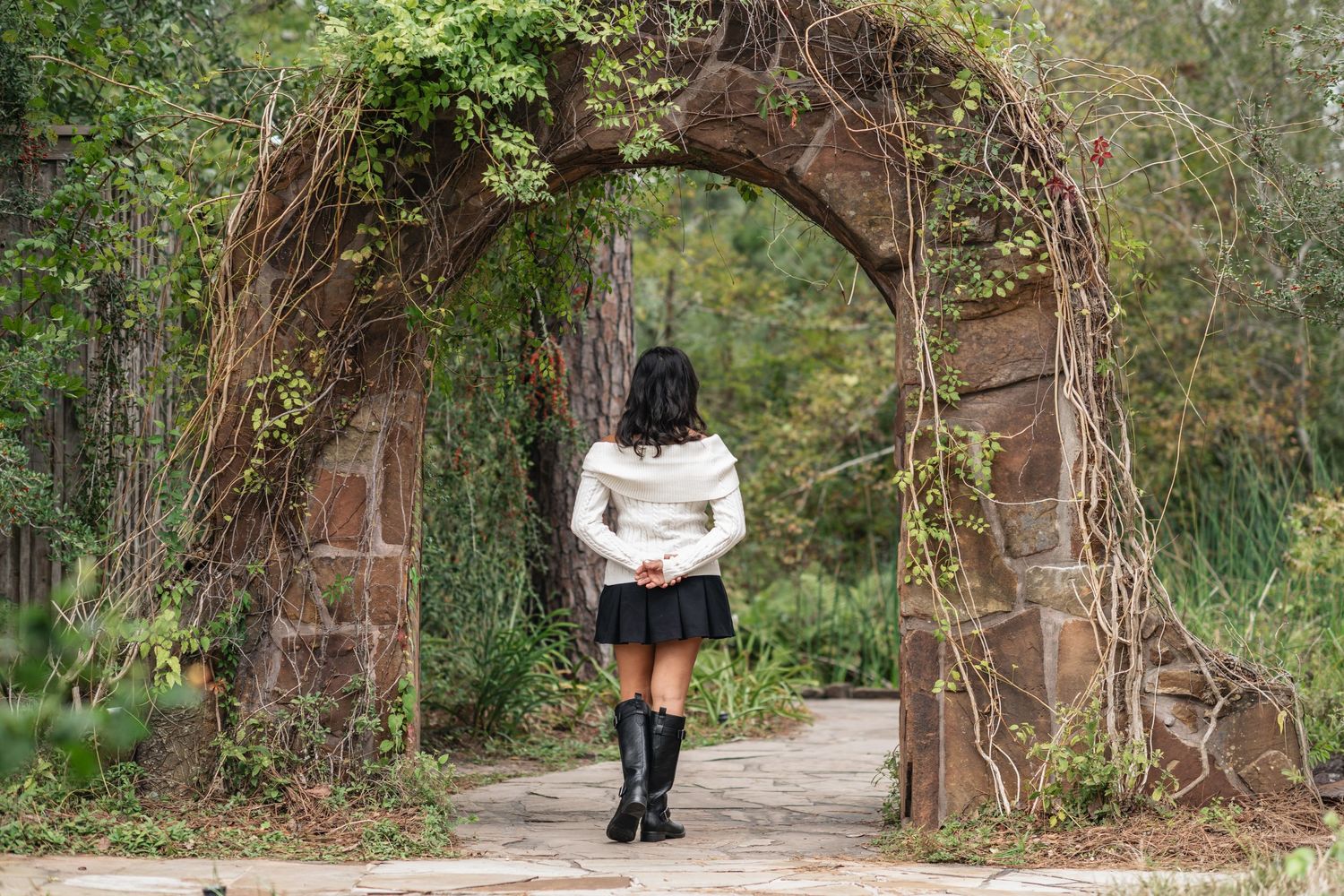 Woman in white sweater and black skirt walking through vine-covered stone archway in garden.