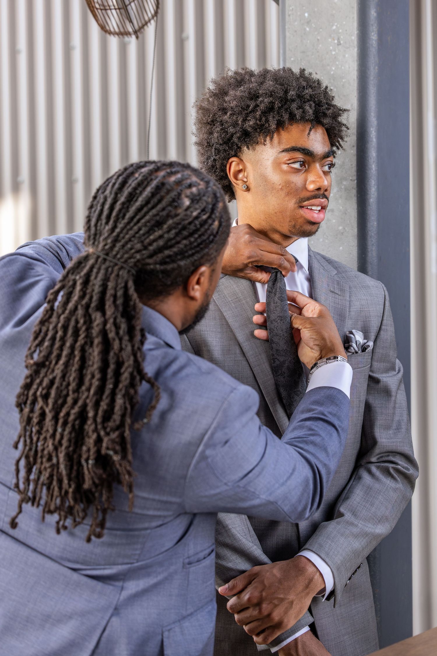 Two Black men in gray suits, one adjusting the other's tie, preparing for a formal event.