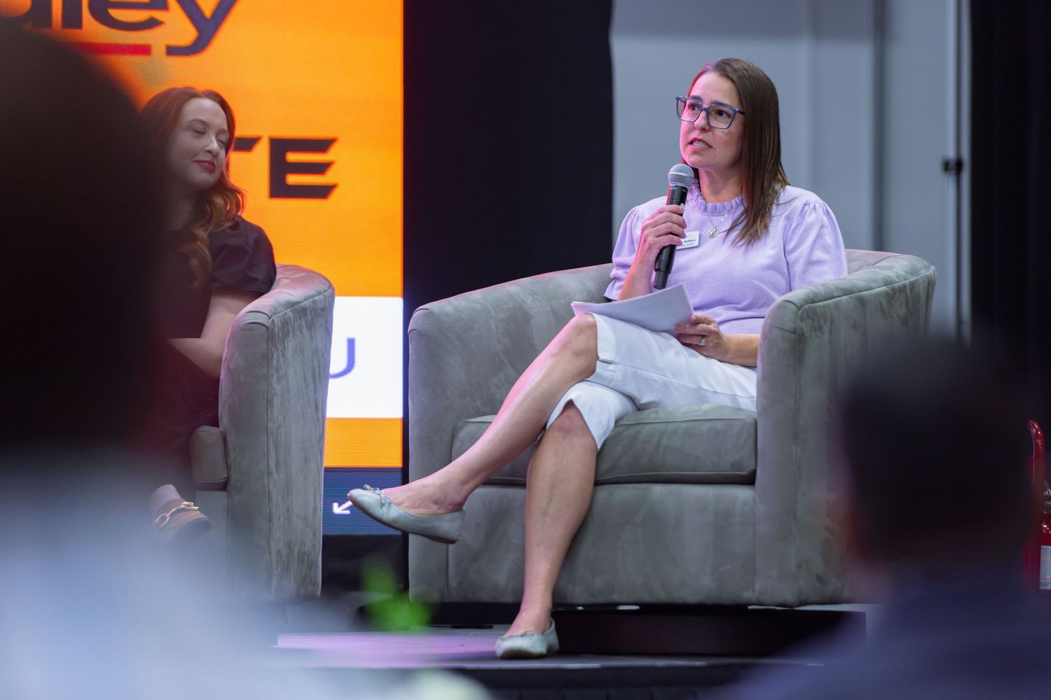 Woman speaking into microphone at panel discussion event, seated in gray chair on stage at conference.