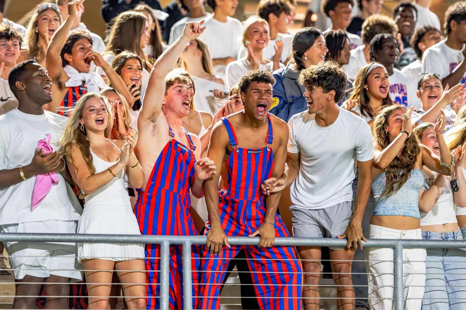Energetic student fans in striped overalls cheer enthusiastically in the bleachers at a football game.