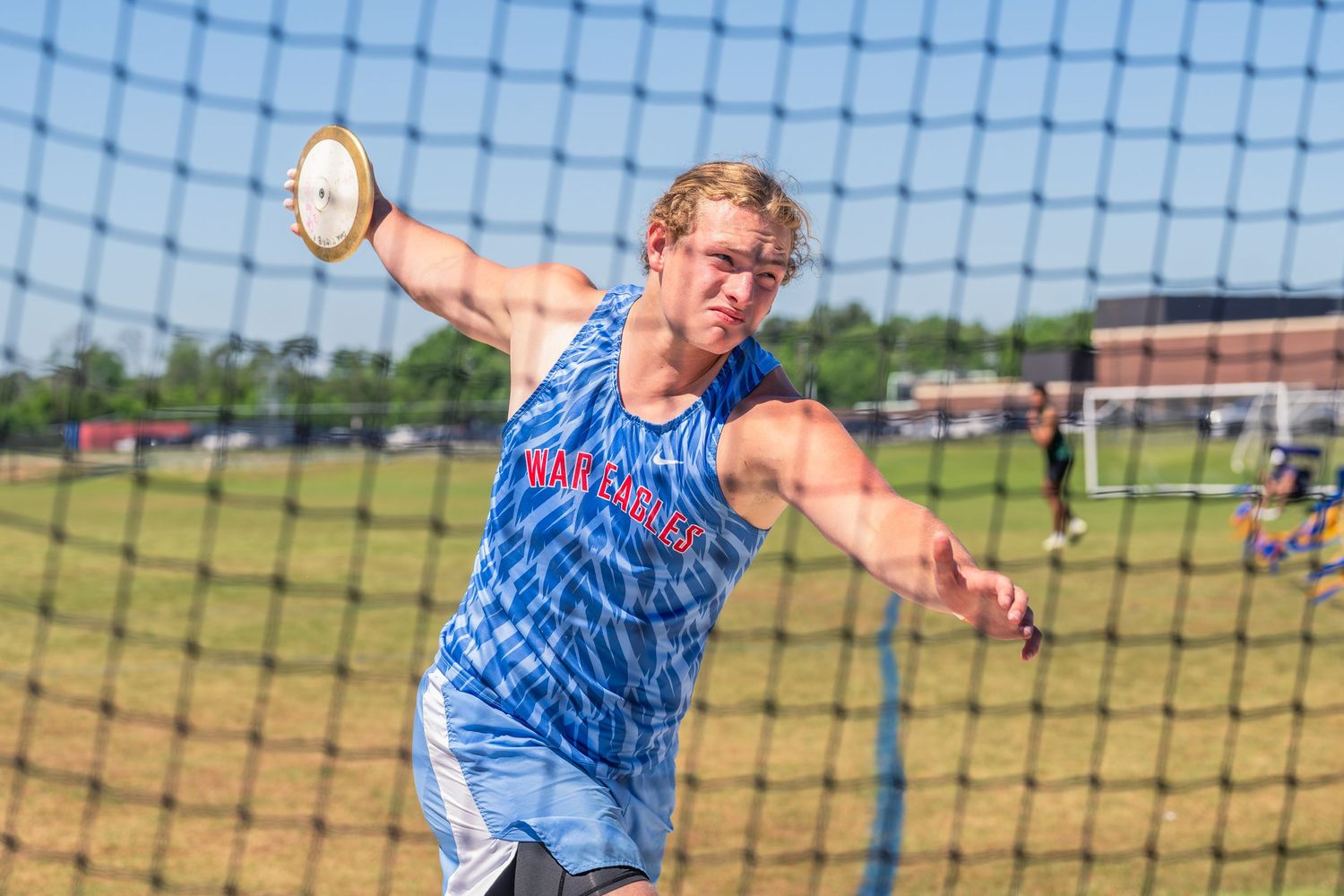 Blonde male athlete in blue uniform throwing discus during a track and field competition.