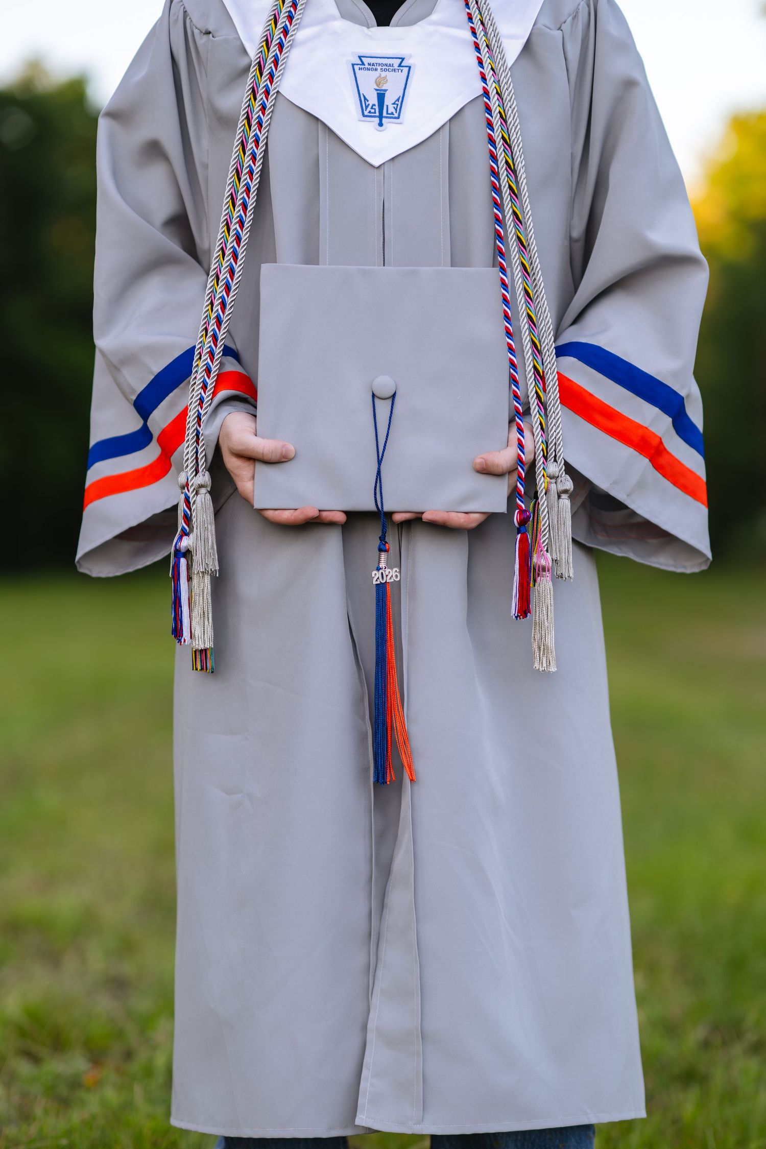 Graduate in gray cap and gown holding diploma with colorful honor cords outdoors.