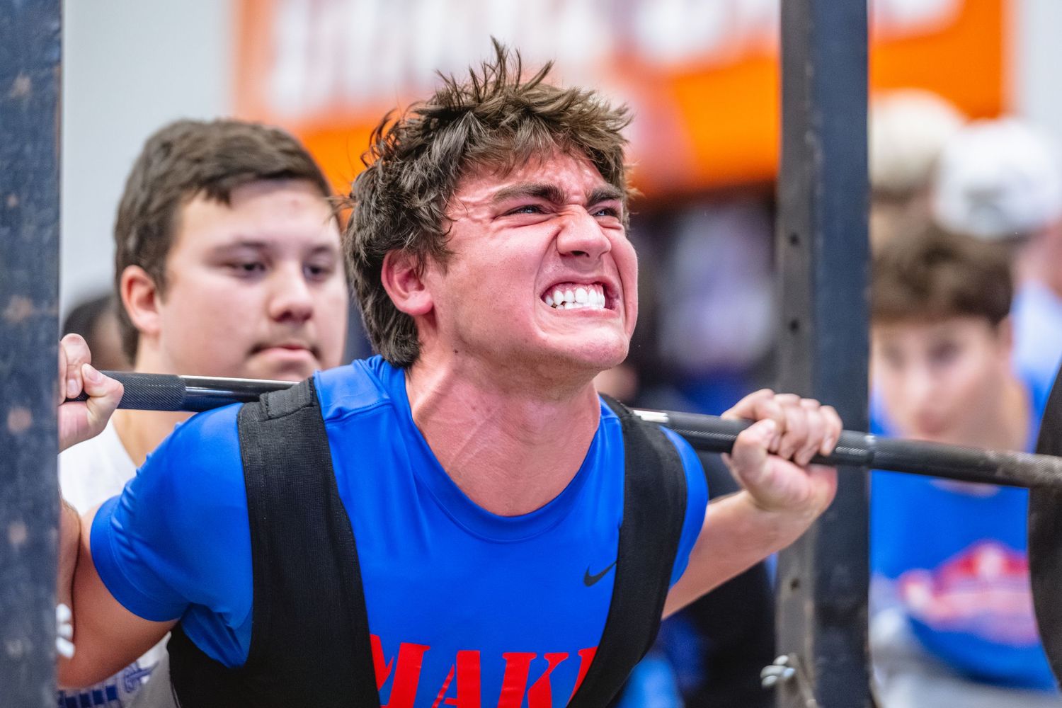 Young athlete grimaces while performing barbell squat in blue Oaks athletic shirt.