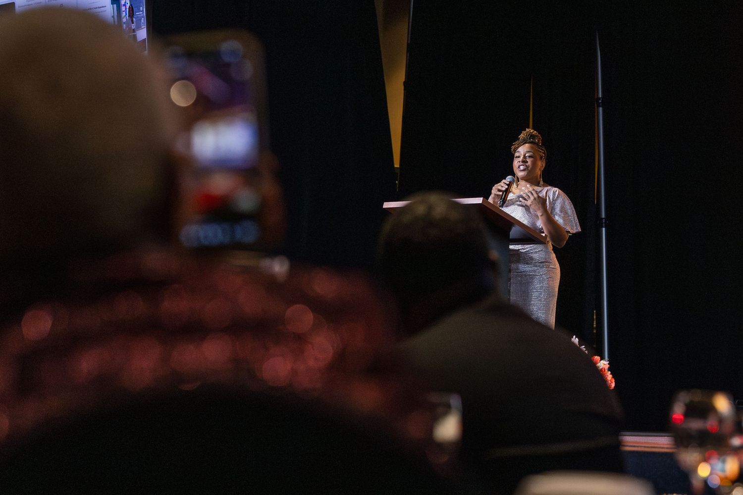 Woman speaks at a podium on a dimly lit stage while audience members record with smartphones in the foreground.