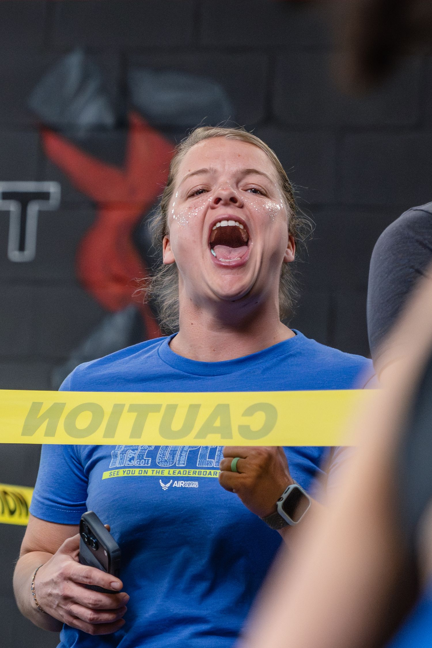 Excited woman in blue shirt cheering behind caution tape at a CrossFit event.