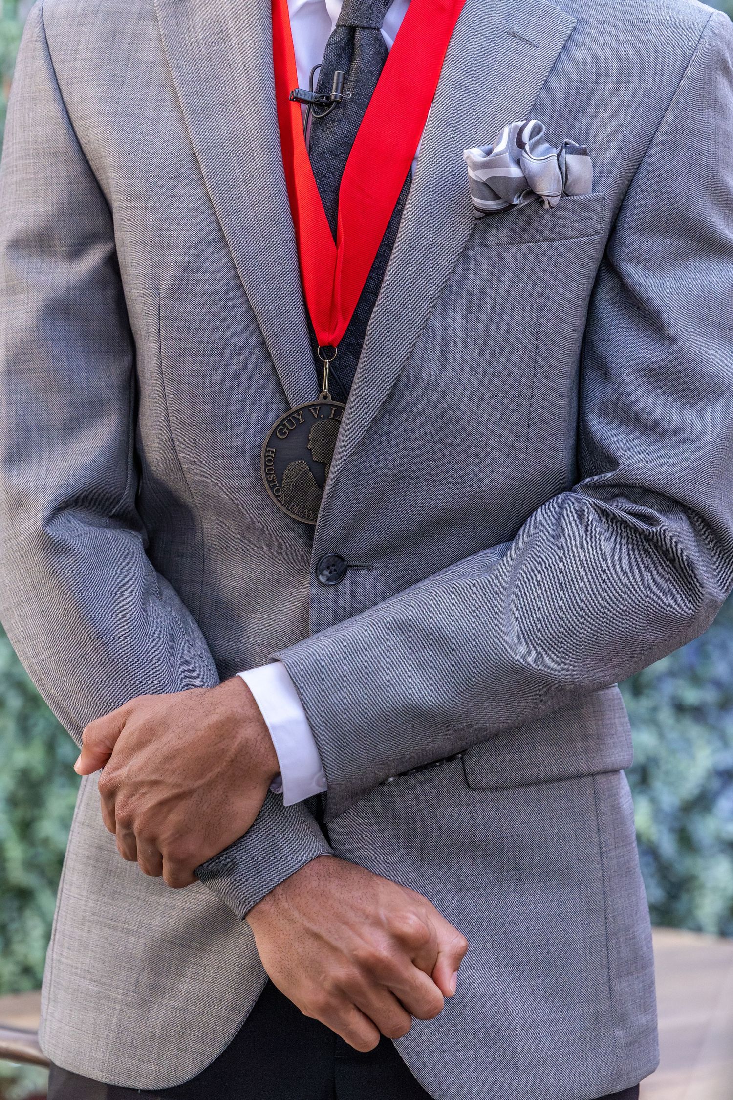Man in gray suit wearing a red ribbon medal with a pocket square, hands clasped, standing outdoors.