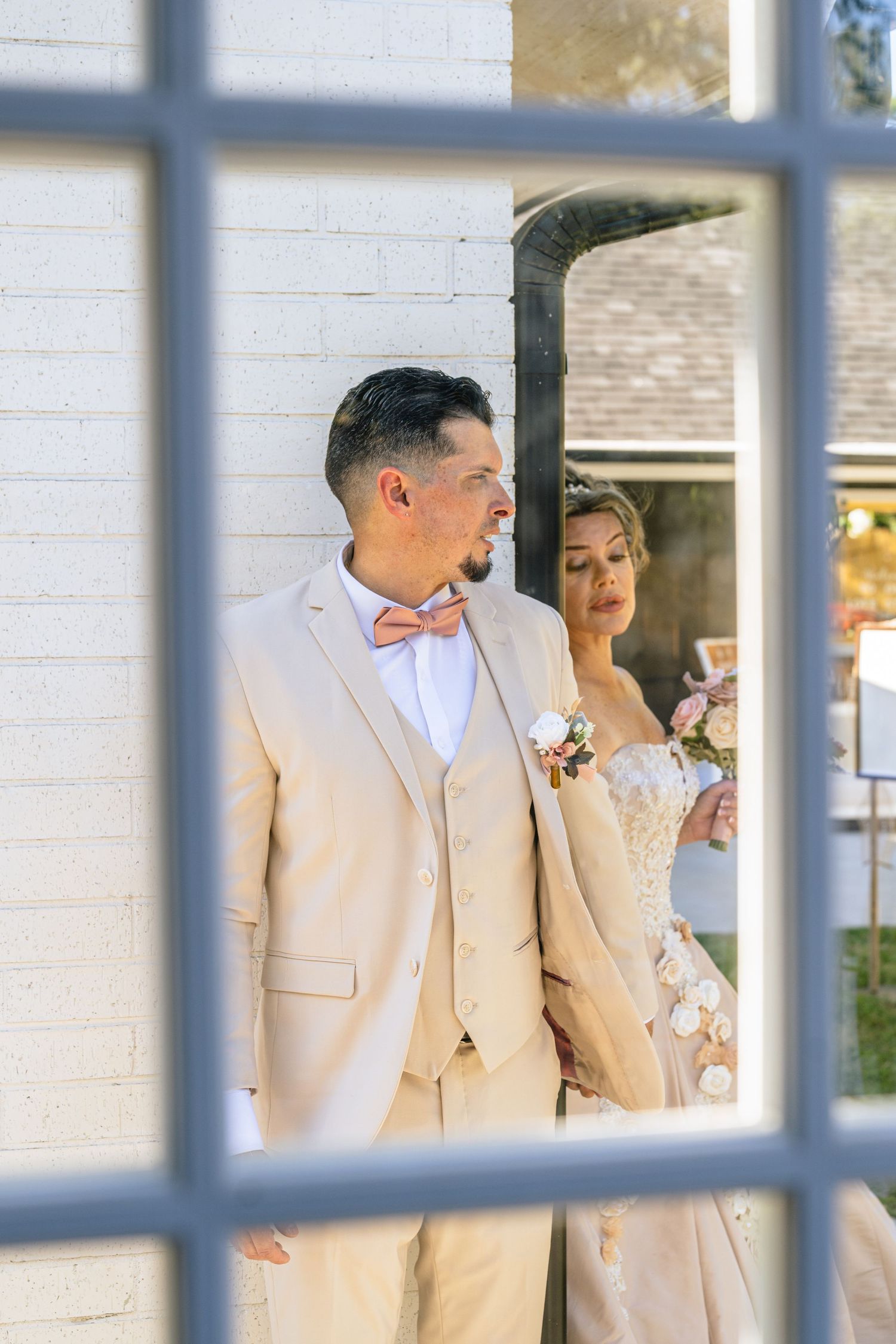 Bride and groom in beige suit with pink bow tie photographed through a window at outdoor wedding venue.