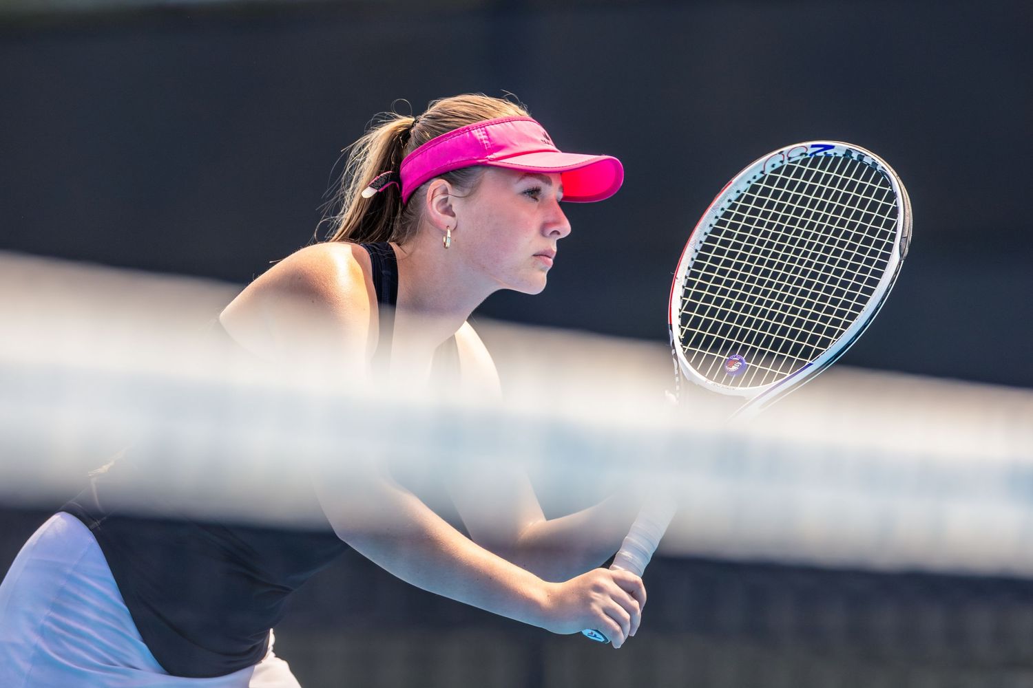 Female tennis player in pink visor and black outfit prepares to return a shot at the net.