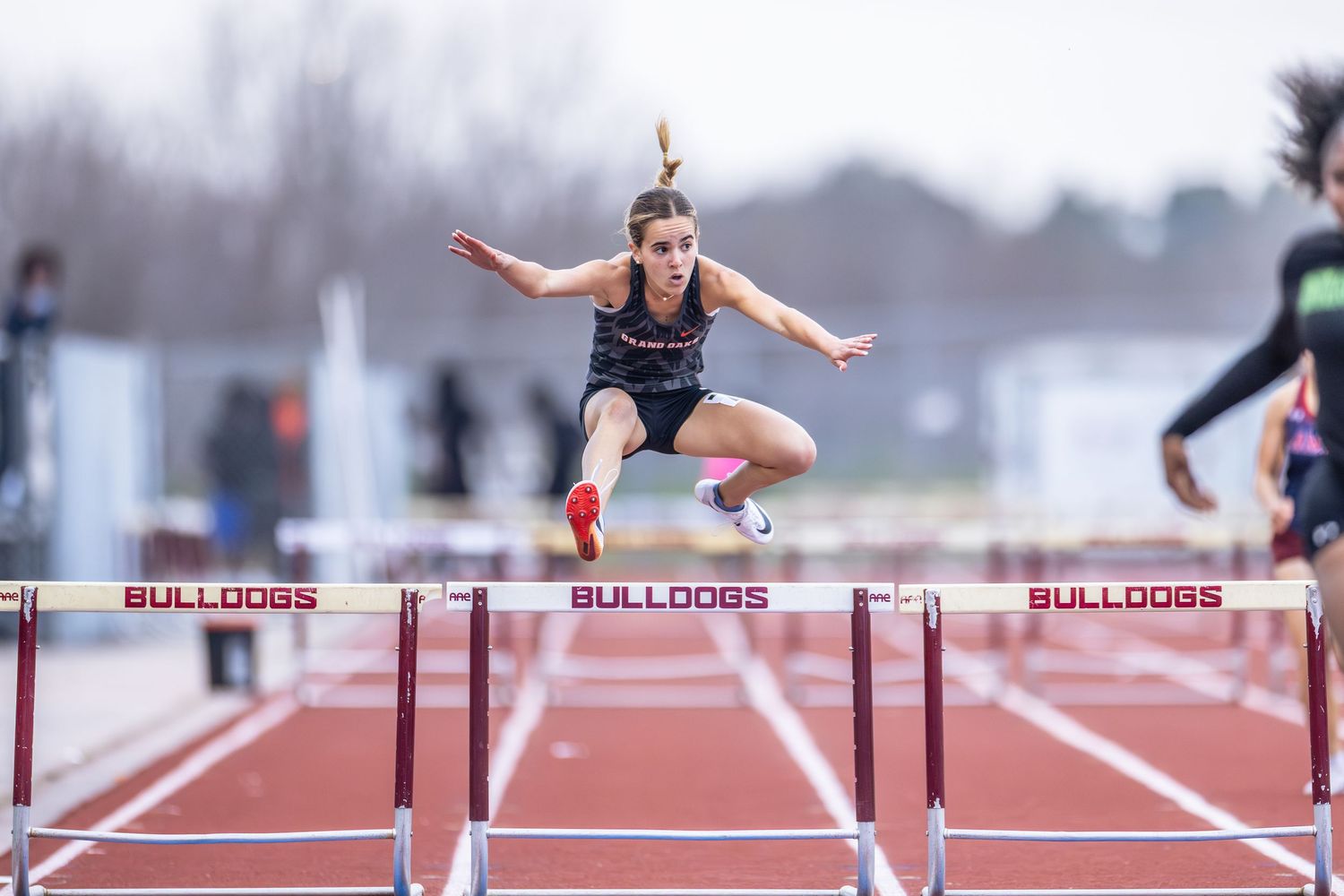 Female hurdler leaping over a hurdle on a Bulldogs track during an outdoor competition.