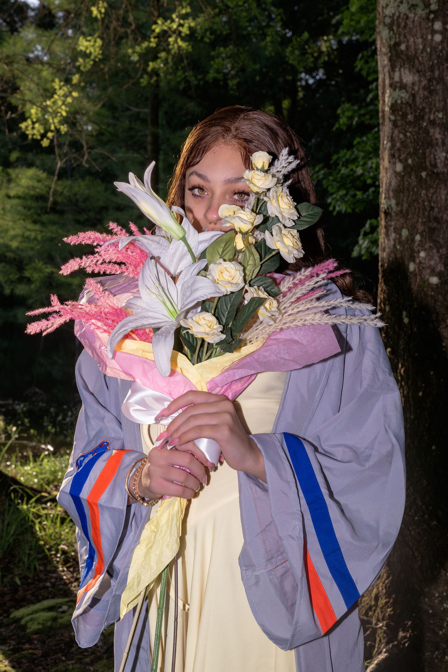 Young woman in graduation gown holding a bouquet of flowers in a sunlit forest.