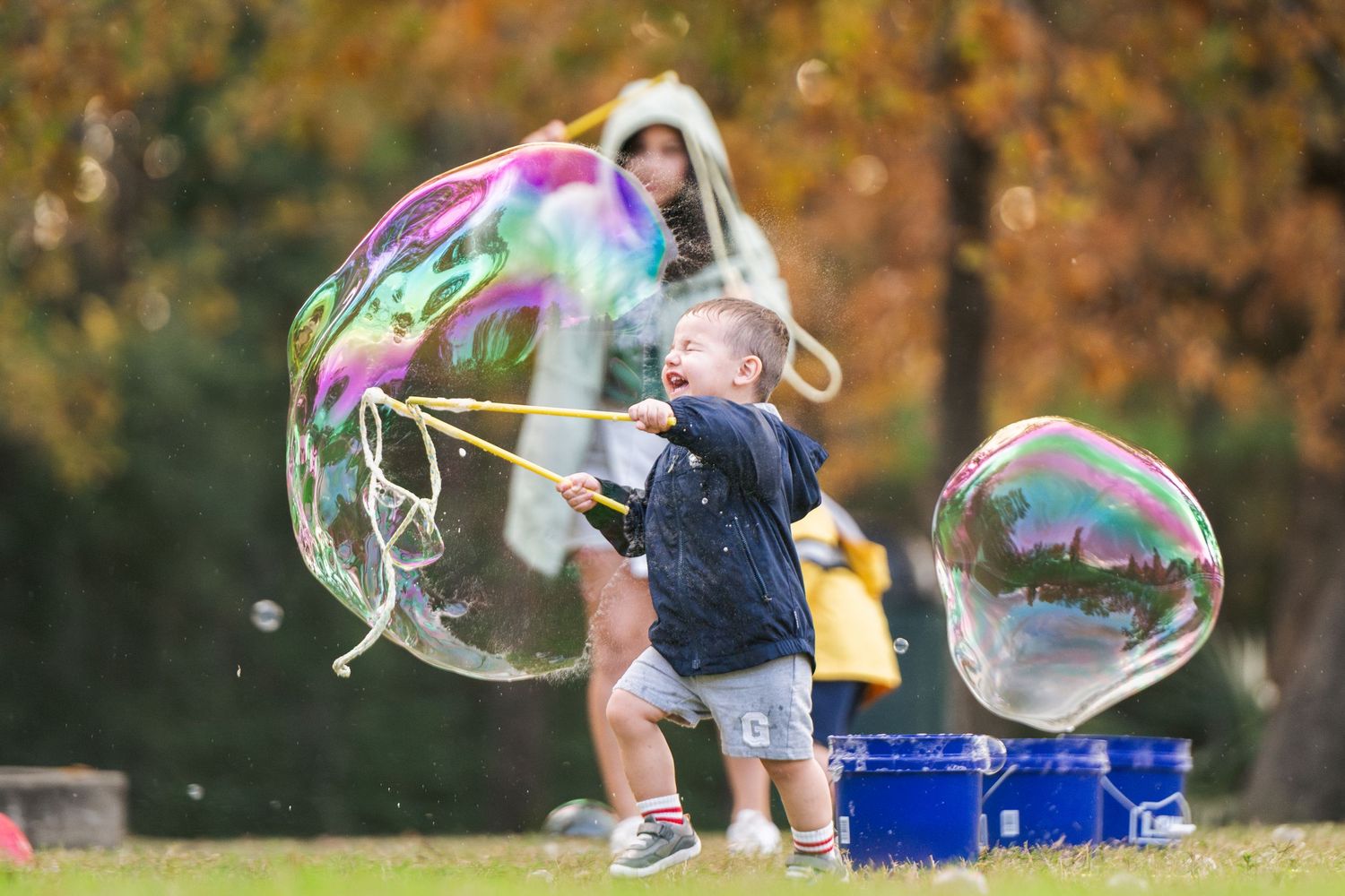 A young child plays with giant soap bubbles in a park during autumn, with colorful trees in the background.