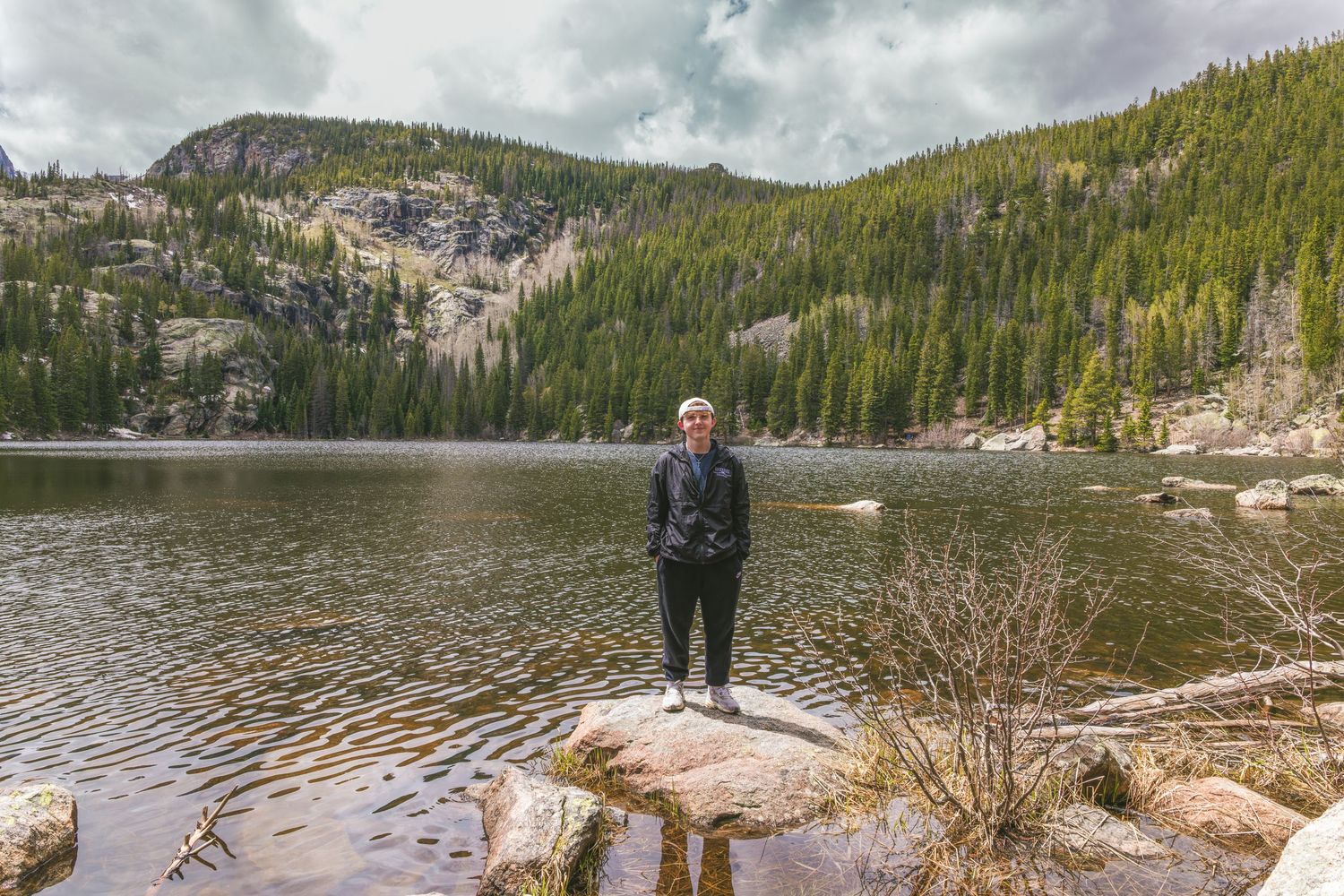 Person standing on rocks beside a serene mountain lake surrounded by pine trees and rocky peaks.