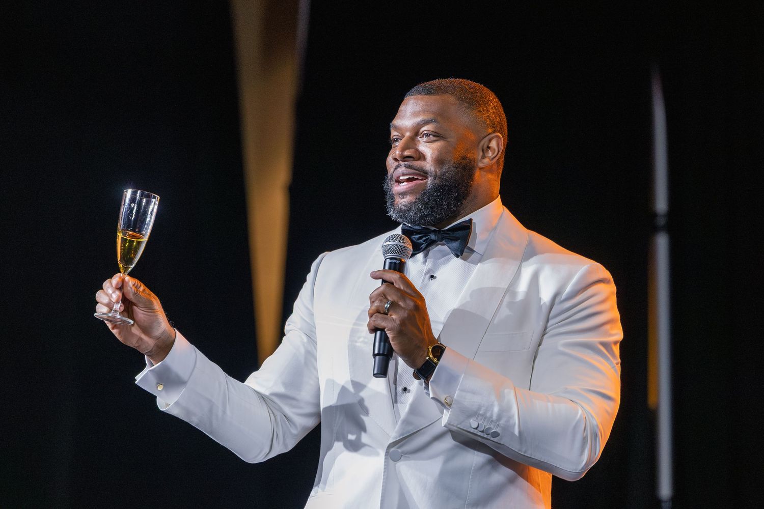 Elegant man in white tuxedo raises champagne glass while holding microphone at a formal gala event.