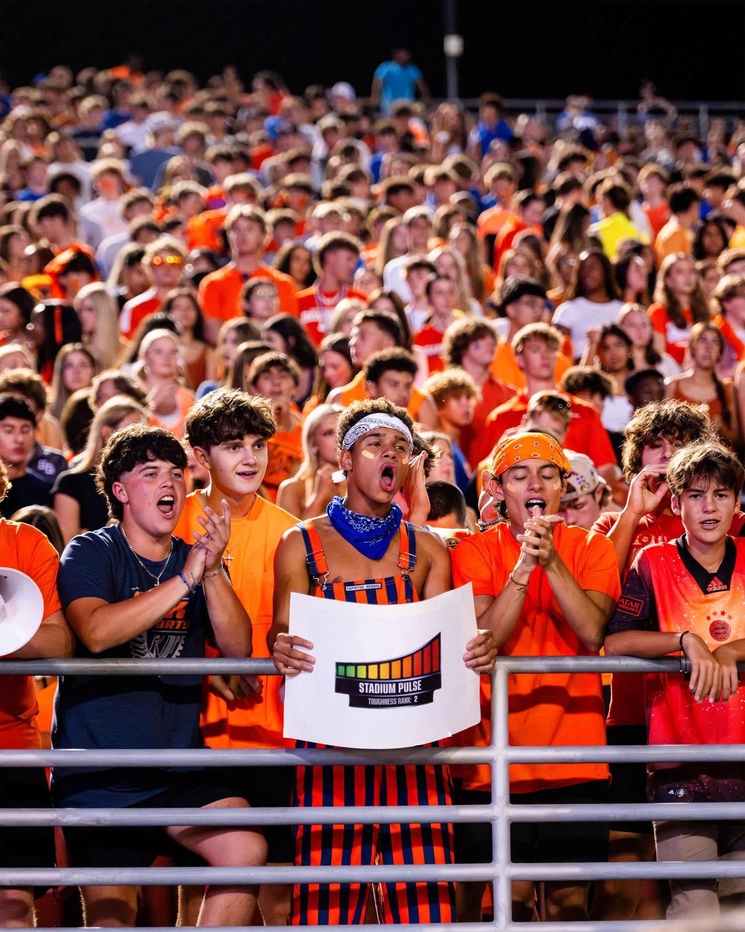 Energetic student section in orange and black cheers loudly at a high school football game.