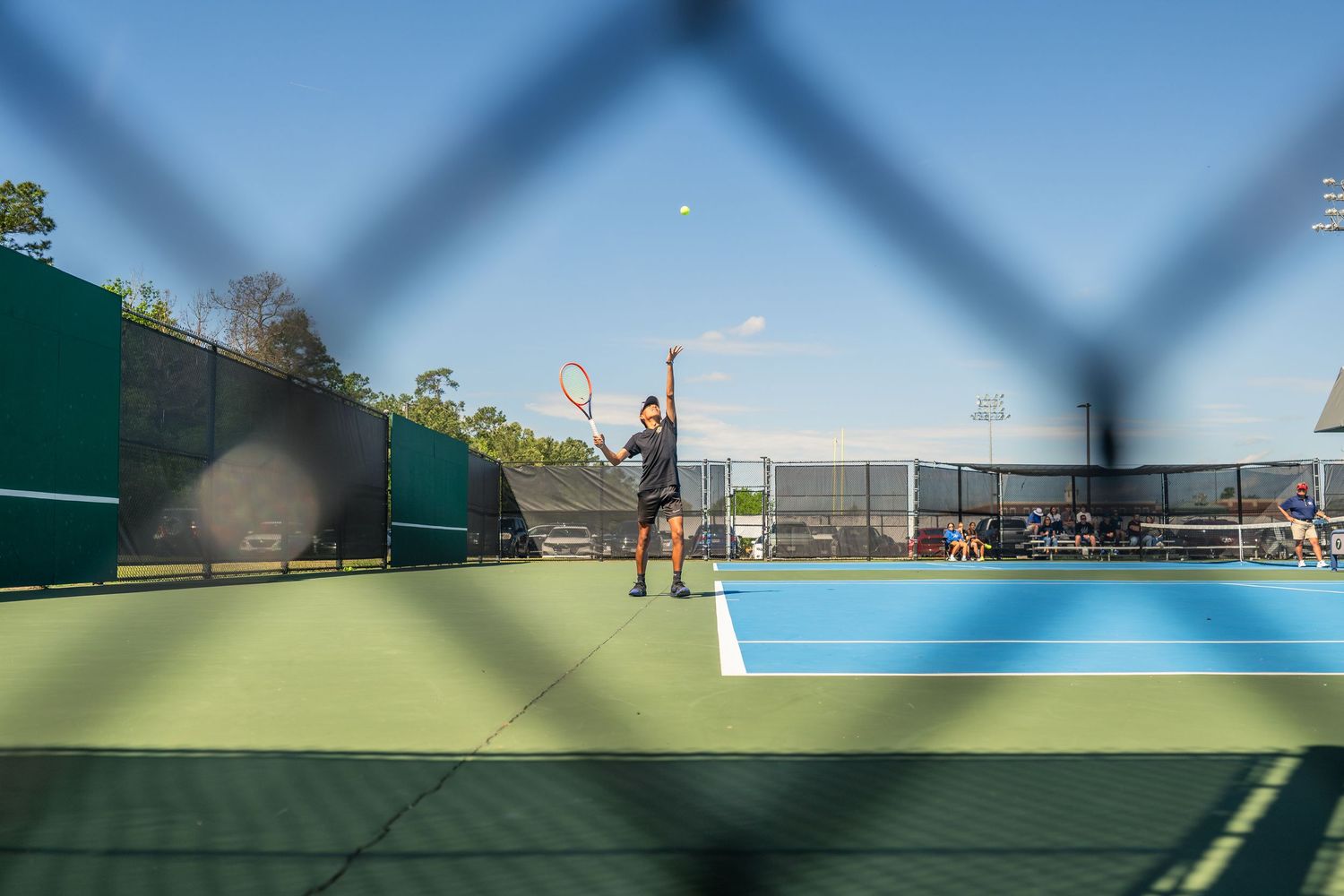 Tennis player serves on a blue outdoor court, photographed through a chain-link fence on a sunny day.