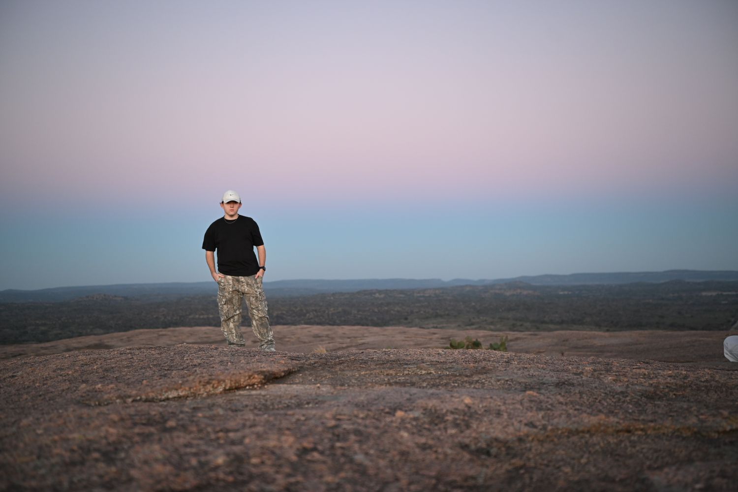 Man standing on a large granite rock at dusk with a vast landscape and pastel sky behind him.