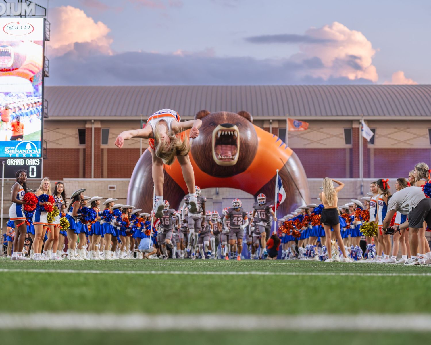 Football team runs through a giant inflatable bear tunnel onto the field at a stadium game night.