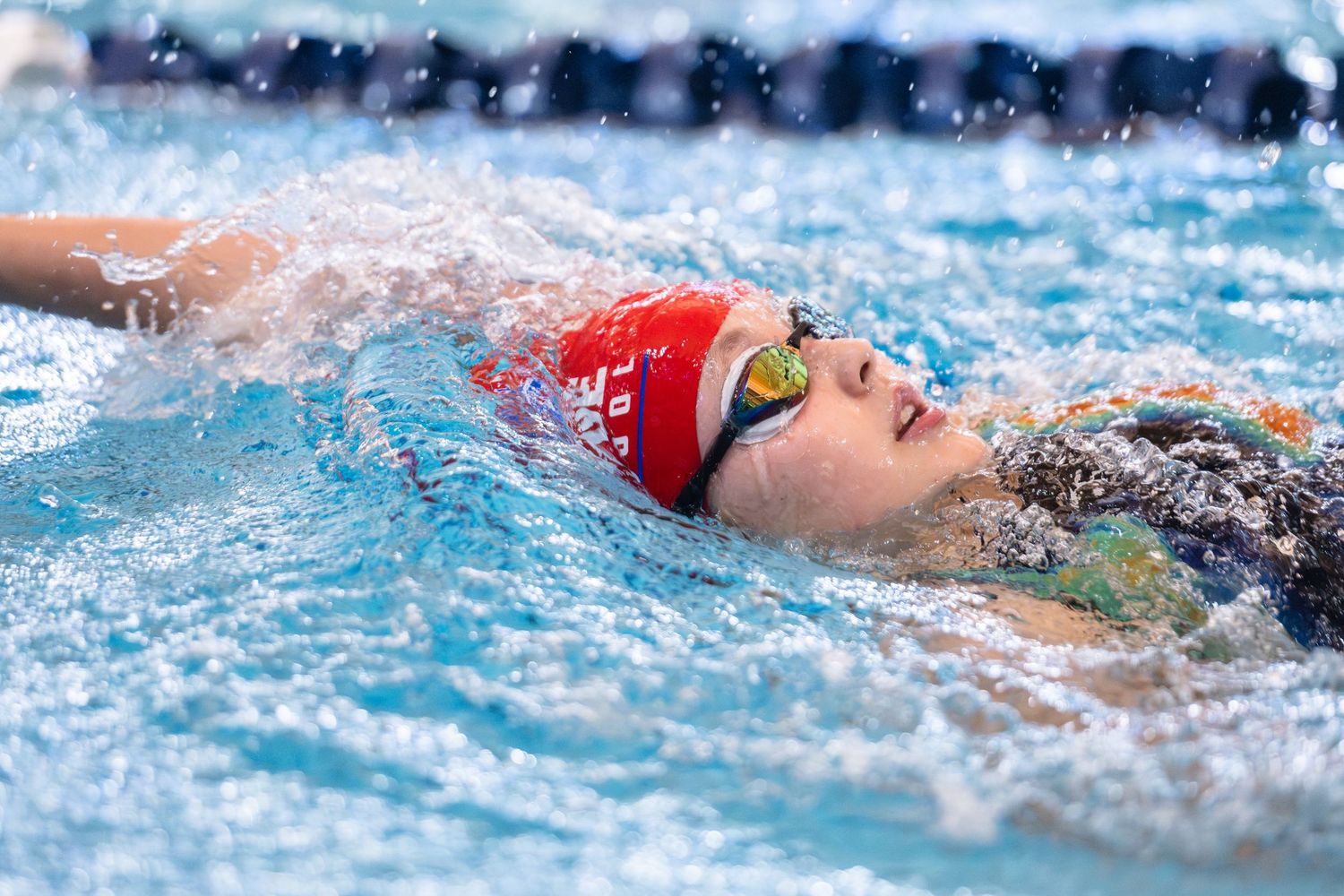 Female swimmer in red cap and goggles performs backstroke in a competitive swimming race.