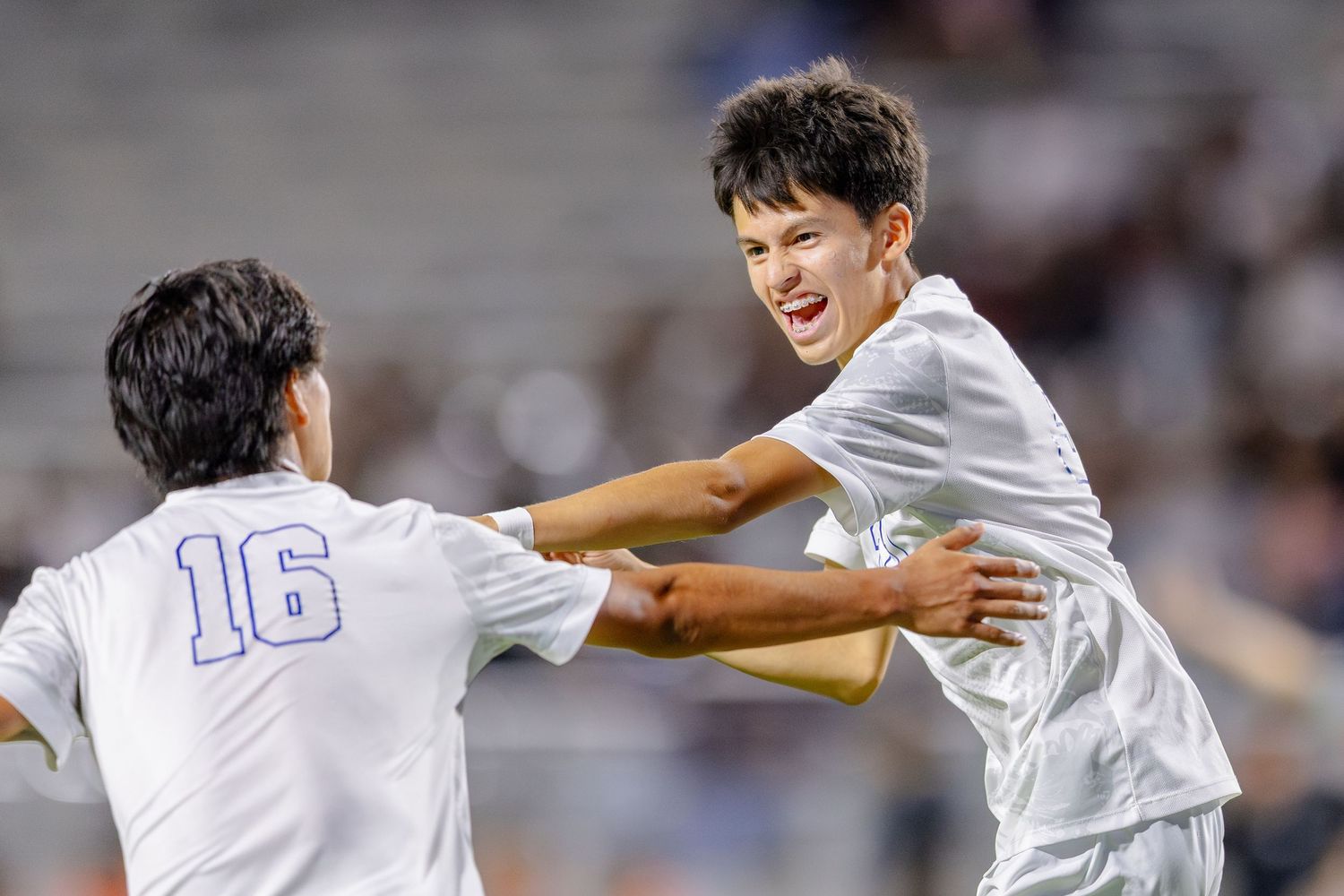 Two soccer players in white uniforms celebrate a goal during a night game.