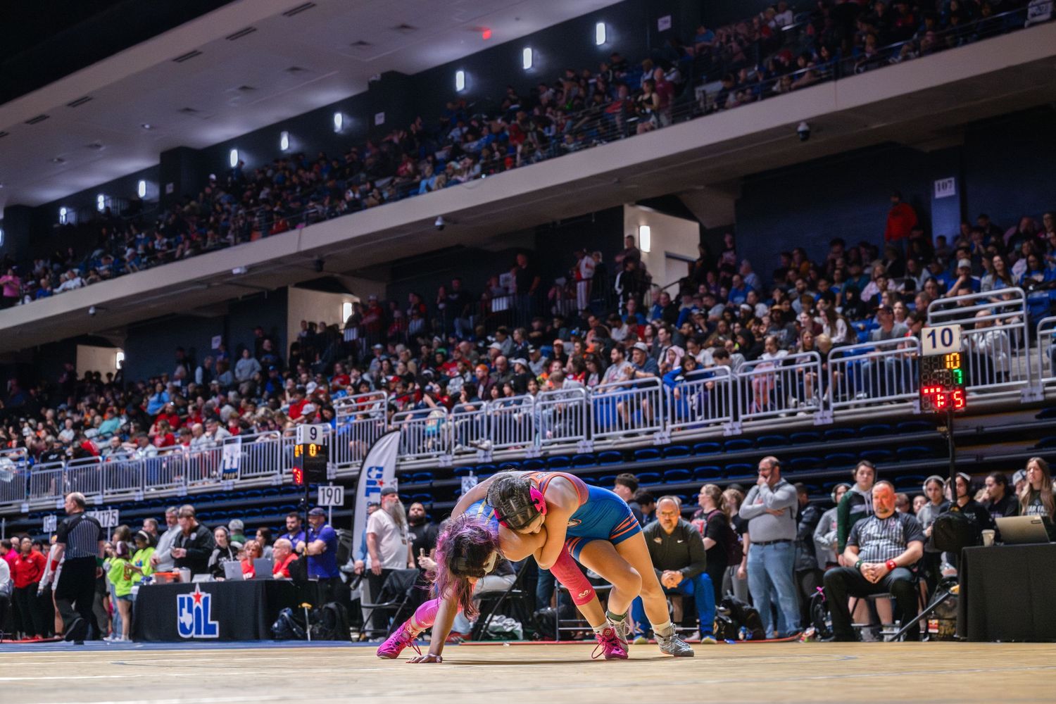 Wrestlers grapple on the mat before a packed arena crowd during a competitive wrestling tournament.