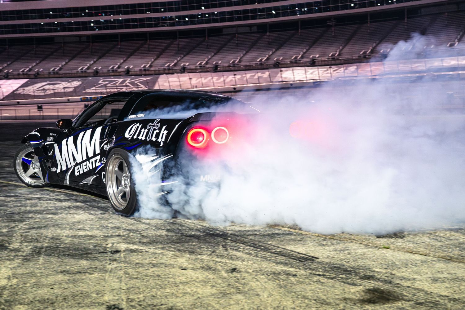 Black drift car performing a smoky burnout at night on a track during an MNM Events clutch competition.