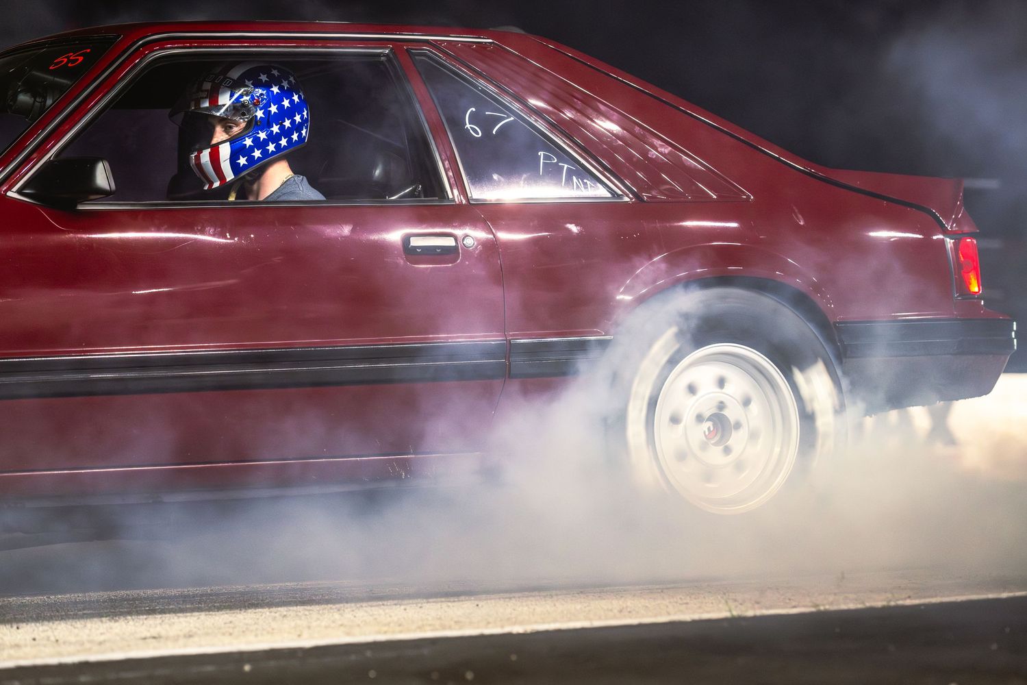 Red Fox Body Mustang performing a burnout with a driver wearing an American flag helmet at night.