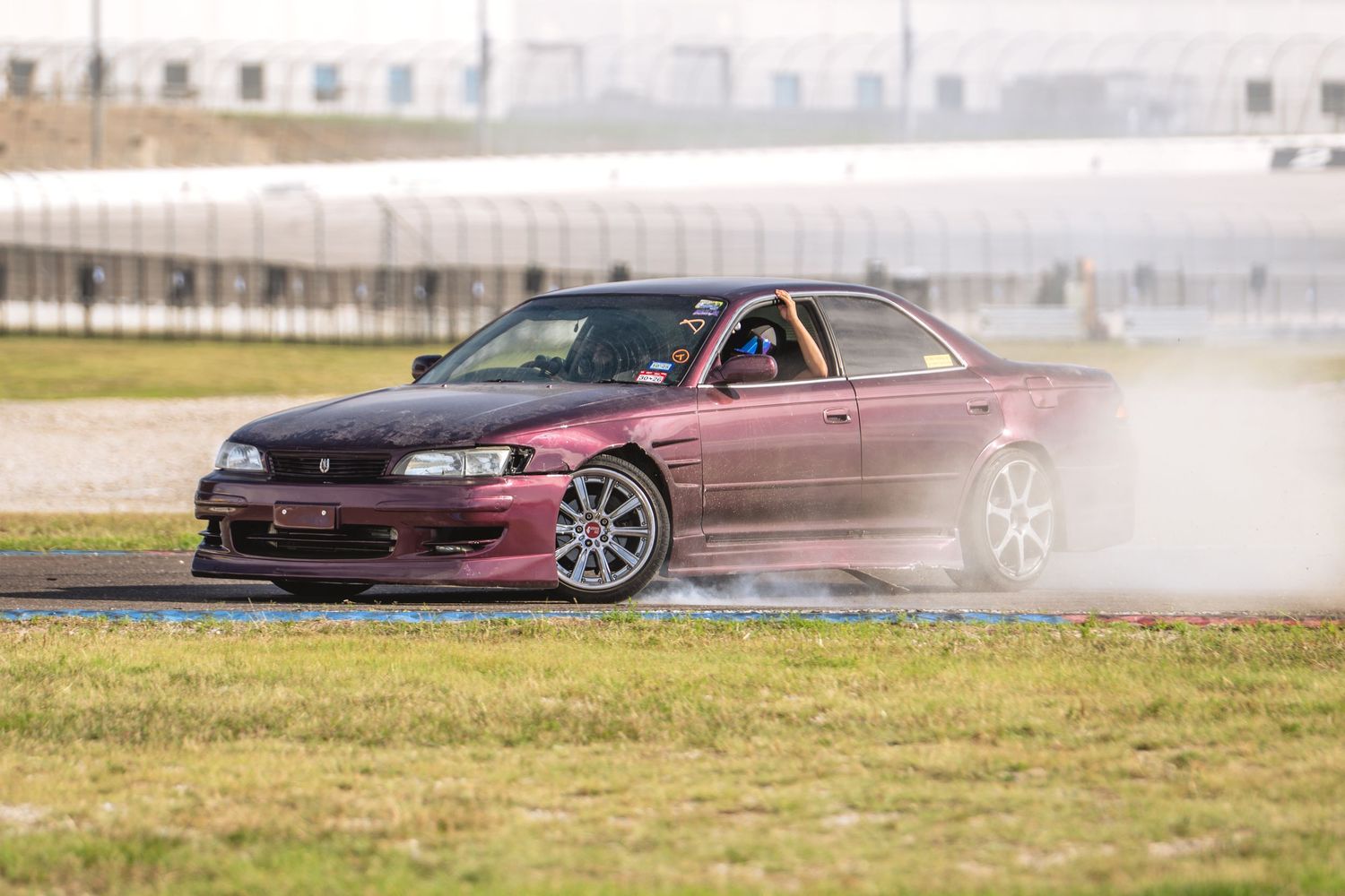Dark maroon Japanese sedan drifting on a track with light tire smoke on a cloudy day.