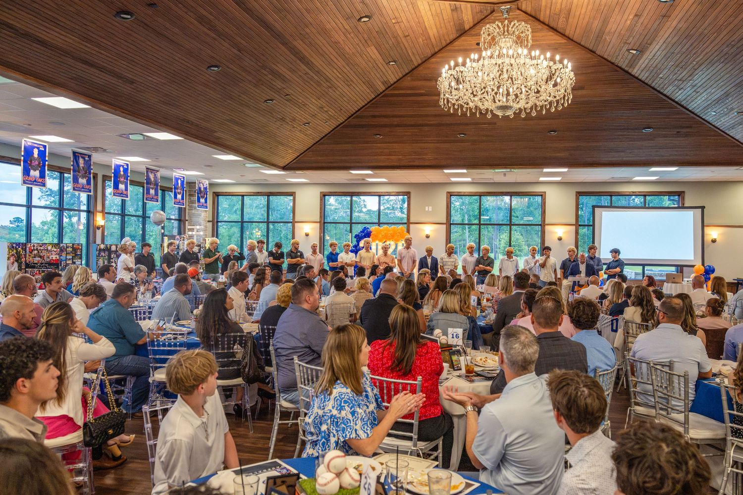 Large banquet hall filled with guests at round tables during a formal event, featuring a chandelier and presentation screen.
