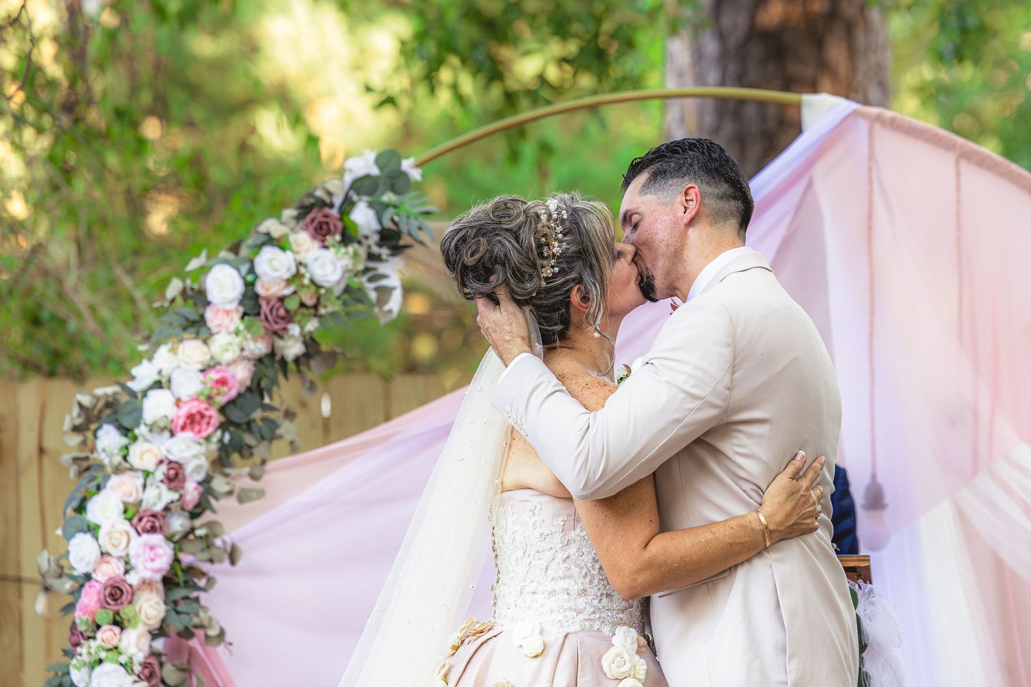 Bride and groom share first kiss at outdoor wedding altar adorned with pink and white floral arch.