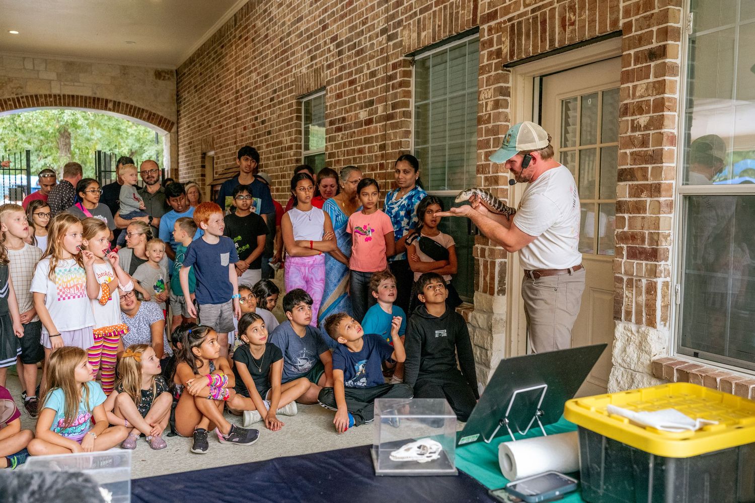 Ranger educator presents wildlife to a large group of children outdoors at a brick building during a nature education program.