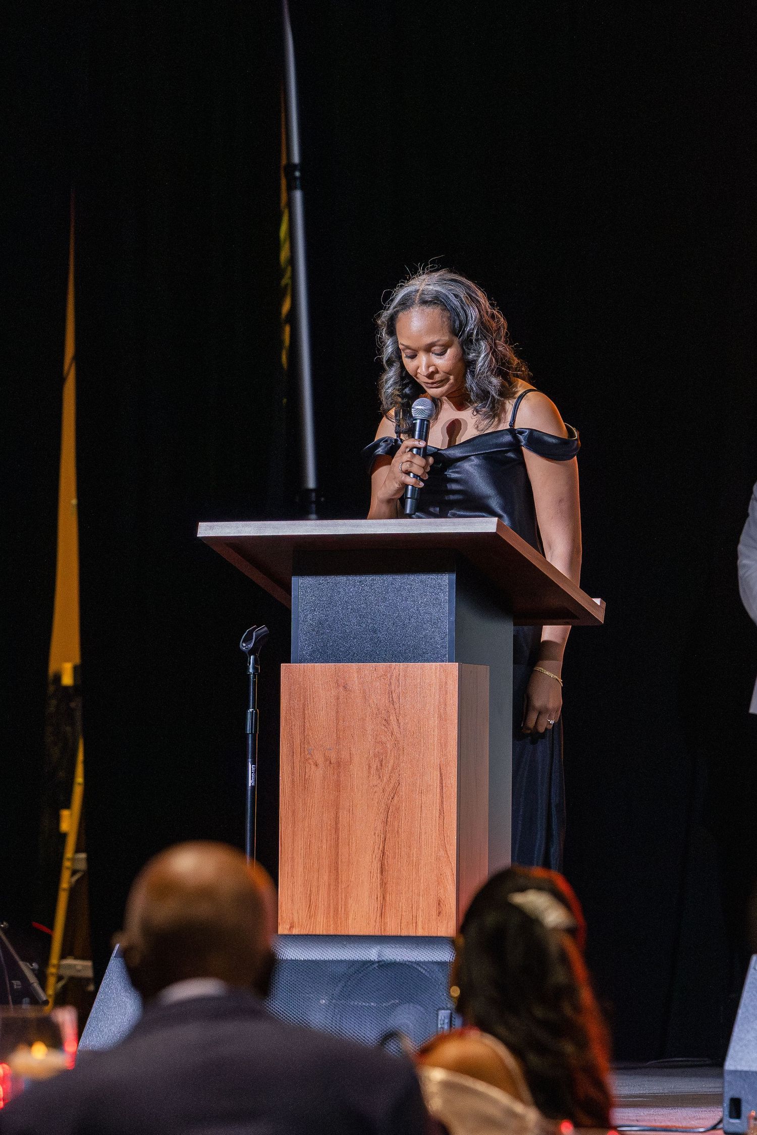 Woman in black off-shoulder gown speaks at a wooden podium on stage before a seated audience at a formal event.