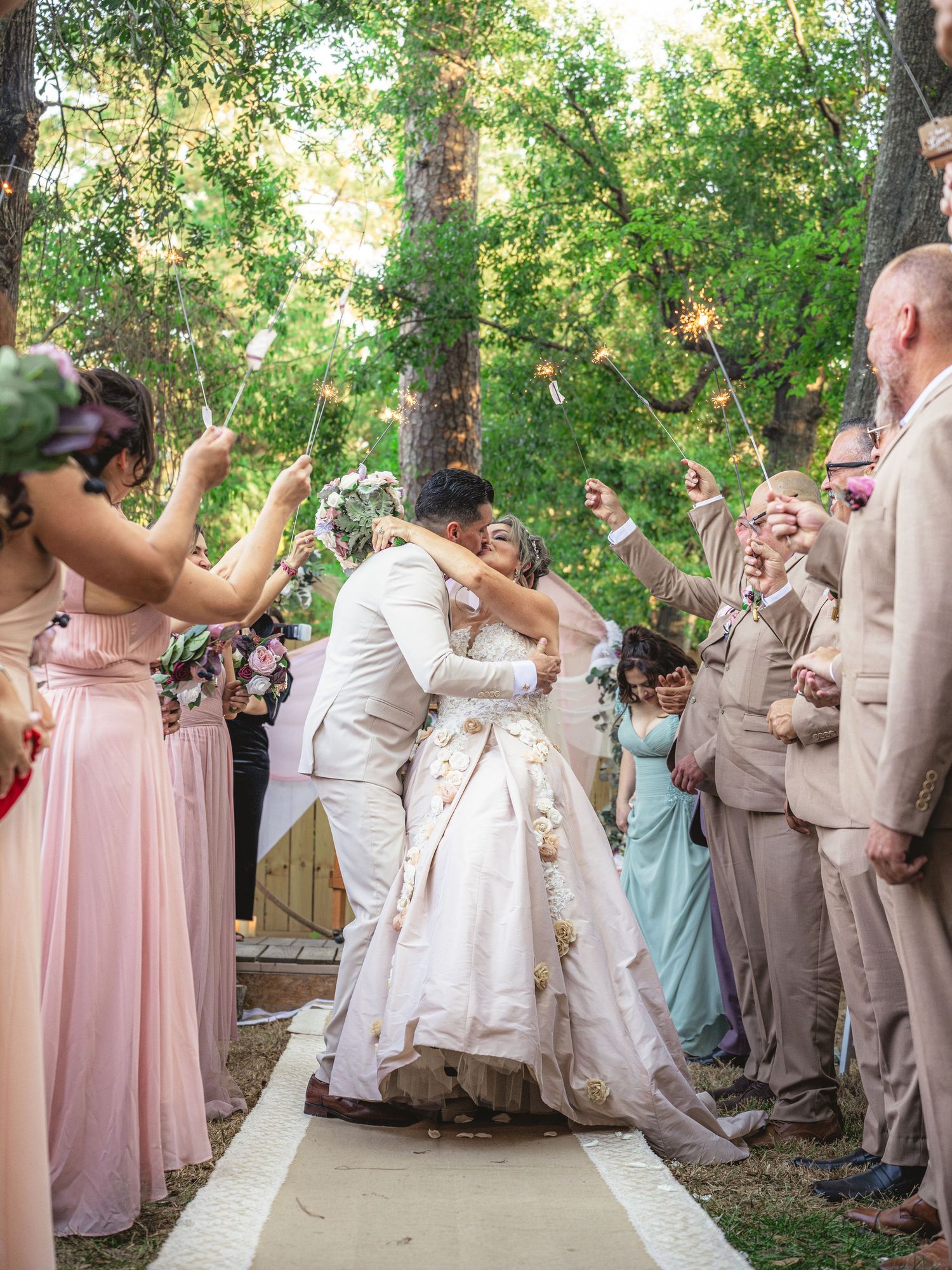 Bride and groom share a kiss during outdoor wedding ceremony while guests hold sparklers lining the aisle.
