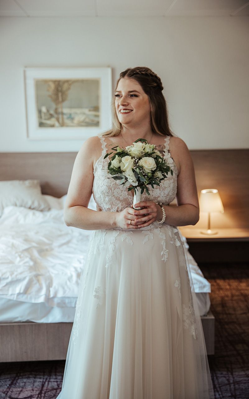 Braut hält Blumenstrauss im Arm und lacht. Im Hotelzimmer.